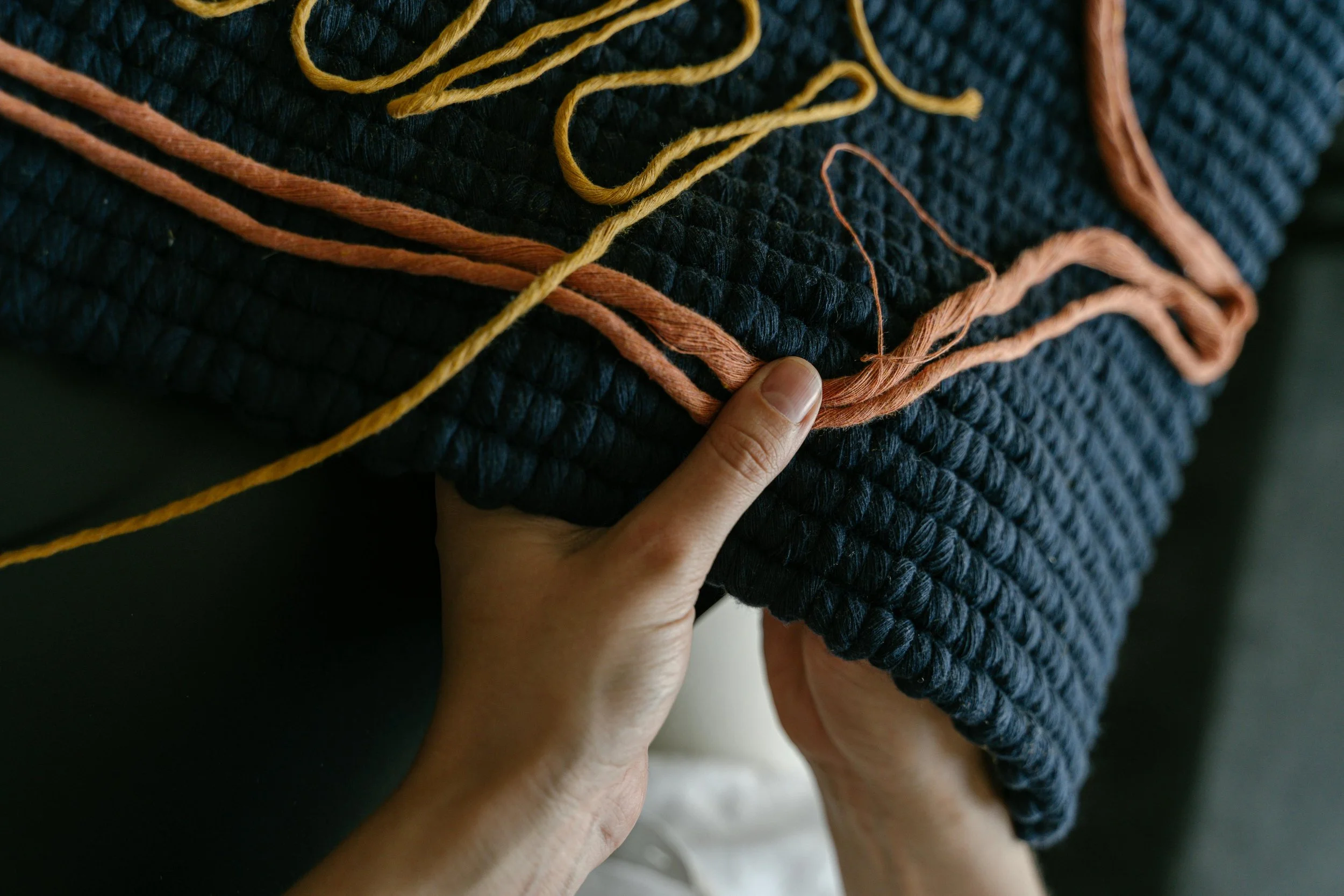 Close-up of a person's hands holding a folded dark blue woven blanket with orange and yellow yarns on top.