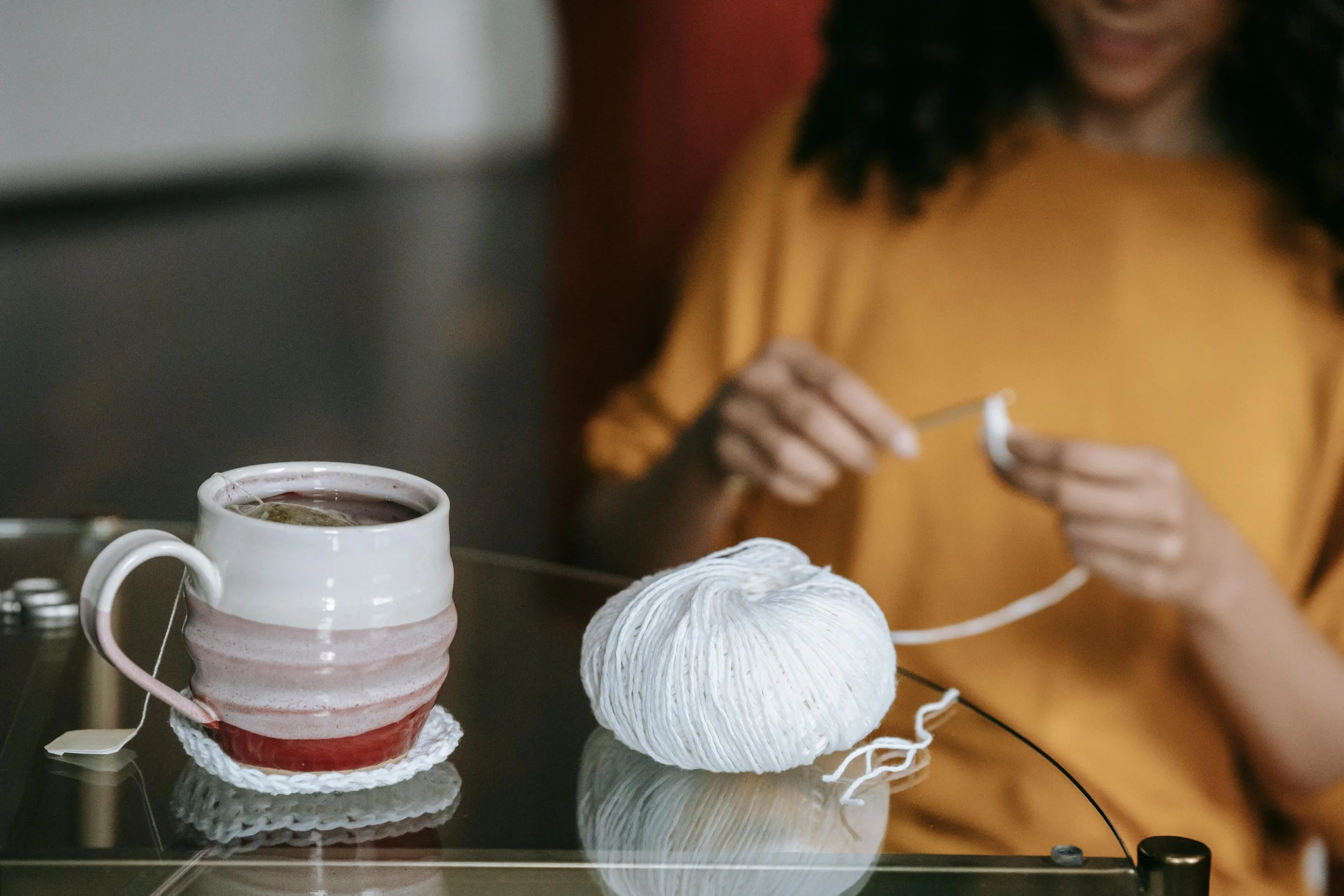 A person in a mustard yellow shirt knitting with white yarn while sitting at a glass table. On the table, there is a mug with a red and white glazed pattern, filled with a hot beverage, and a ball of white yarn.