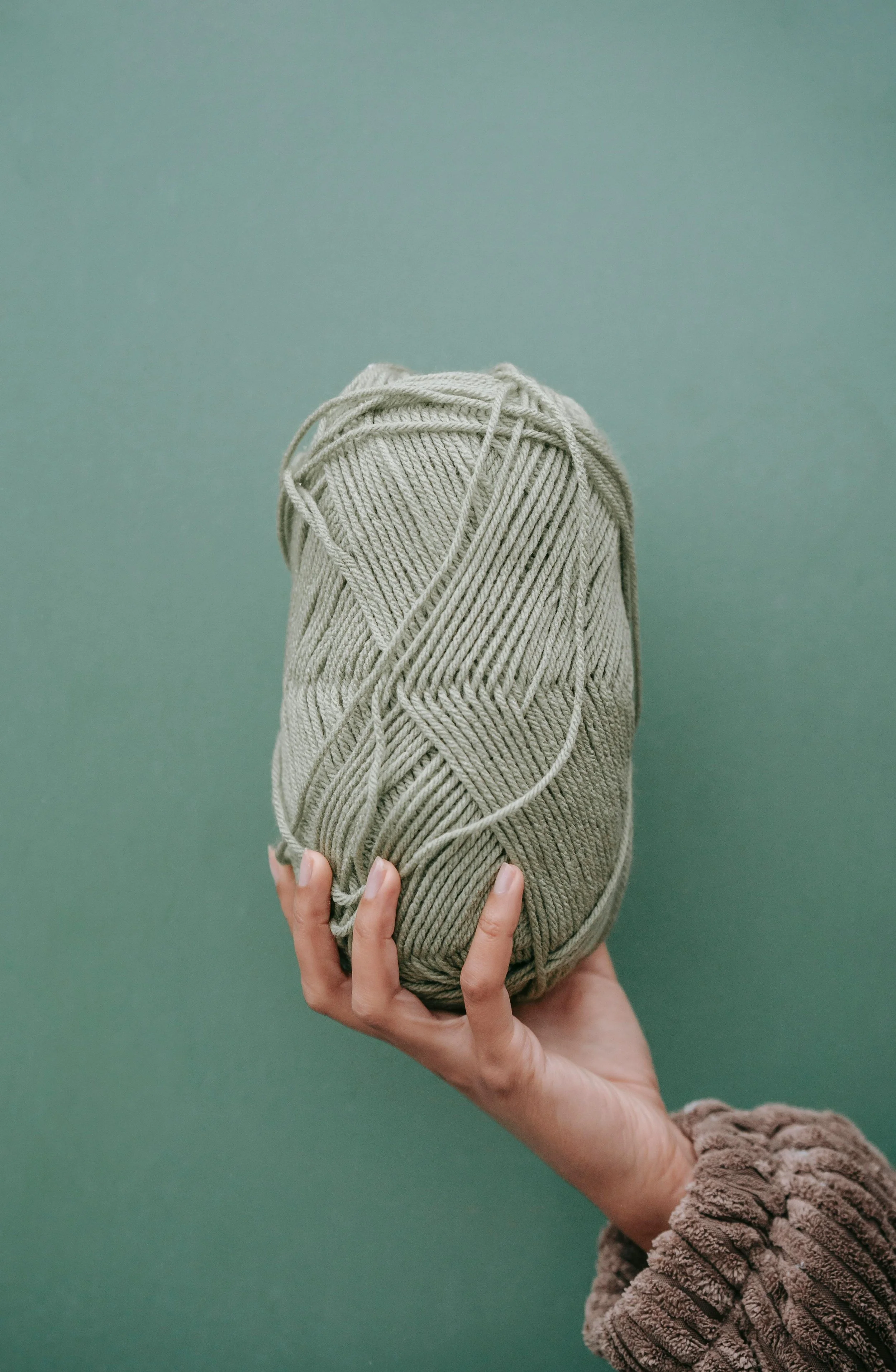 Hand holding a large ball of beige yarn against a green background.