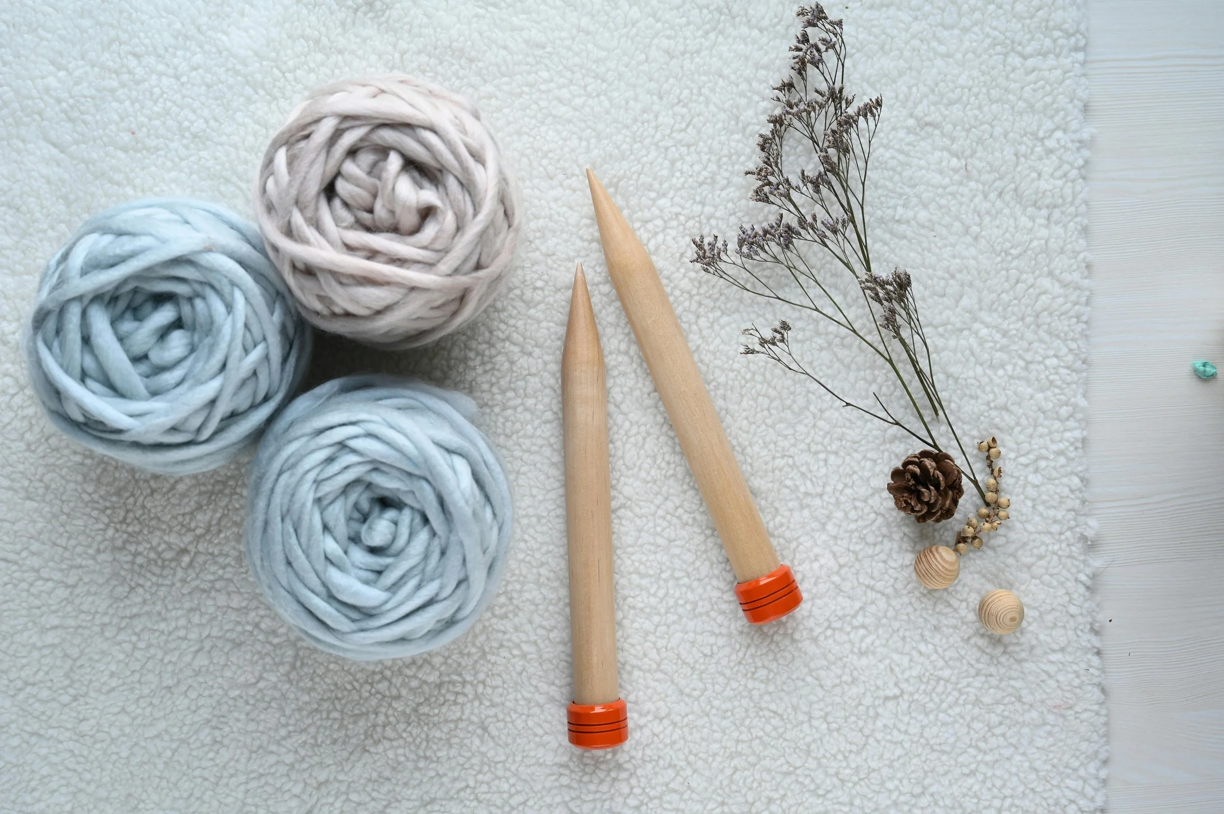 Three balls of chunky yarn in light blue and beige colors, two wooden knitting needles with orange caps, a dried branch with small flowers, a pine cone, and two wooden beads are arranged on a soft textured white surface.