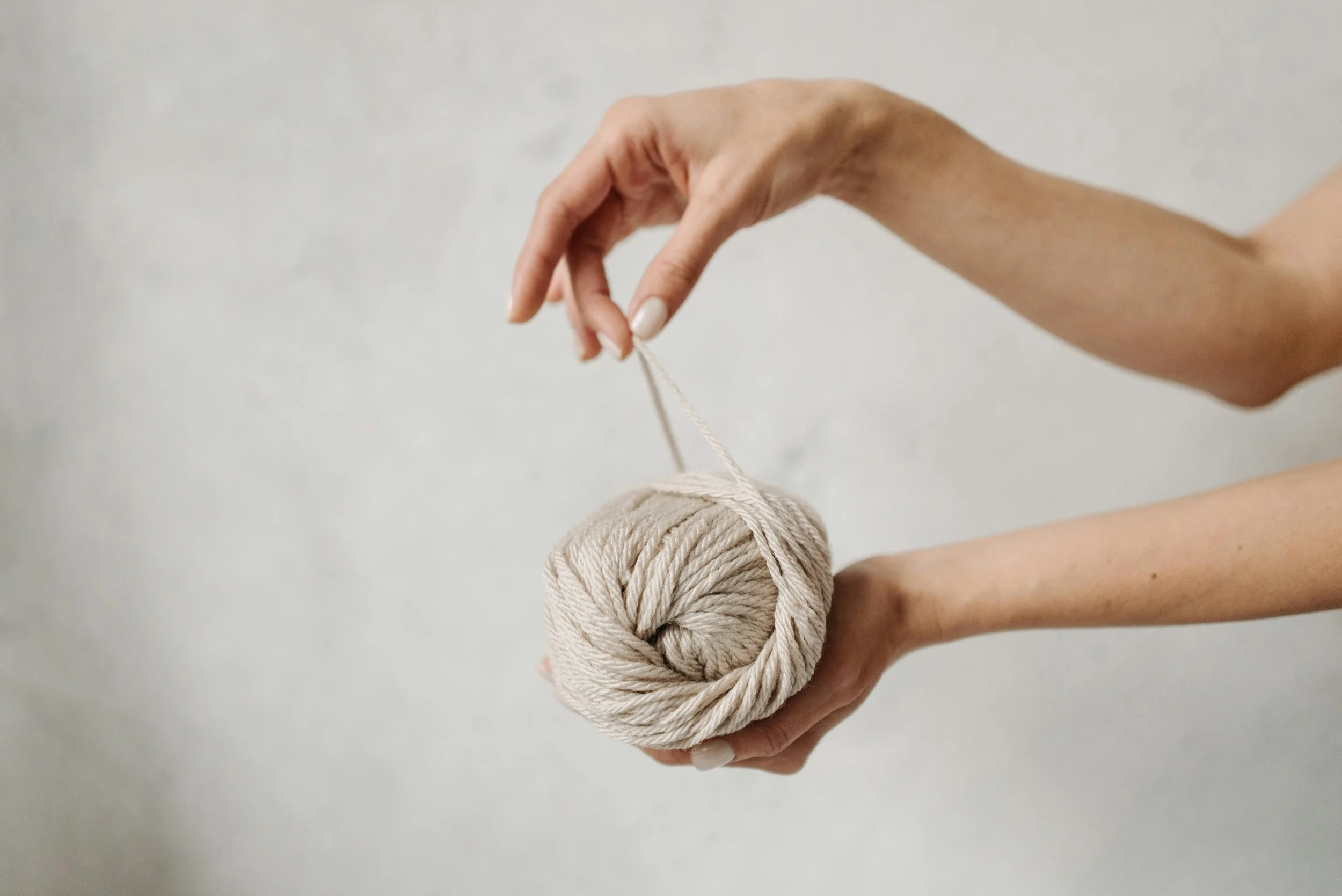 Person holds a ball of beige yarn with one hand and threads a piece of yarn into a knitting needle with the other hand.