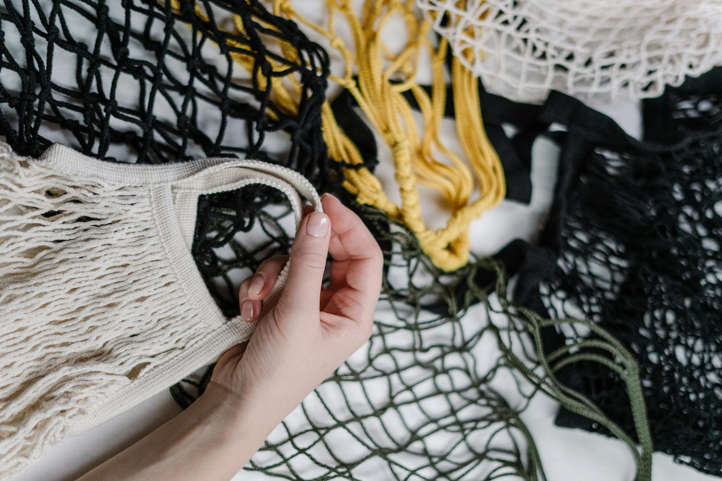 A hand holding a beige netted bag among other colorful mesh bags in black, yellow, and green.