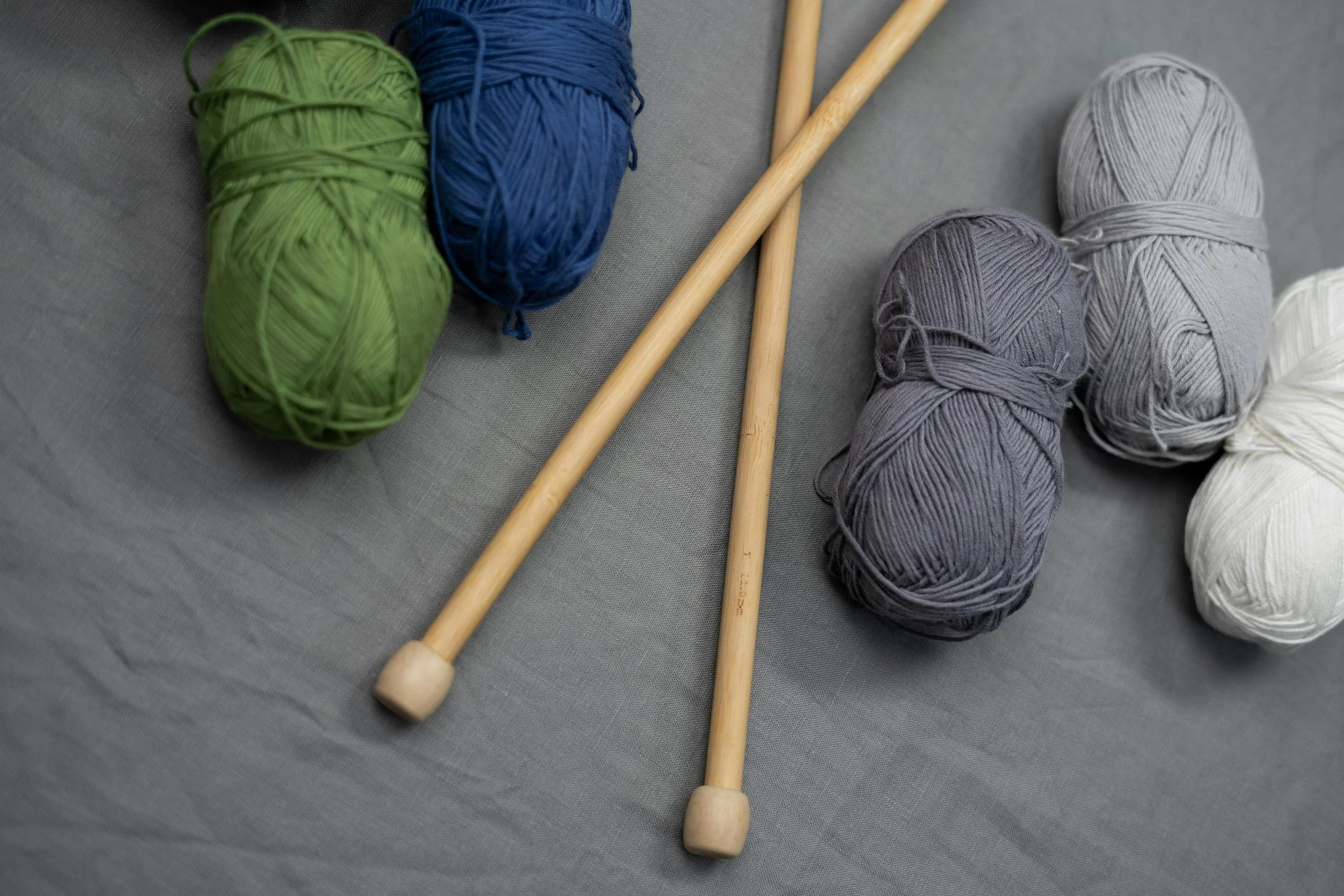 Skeins of yarn in green, blue, gray, white, and purple, along with two wooden knitting needles on a gray fabric surface.