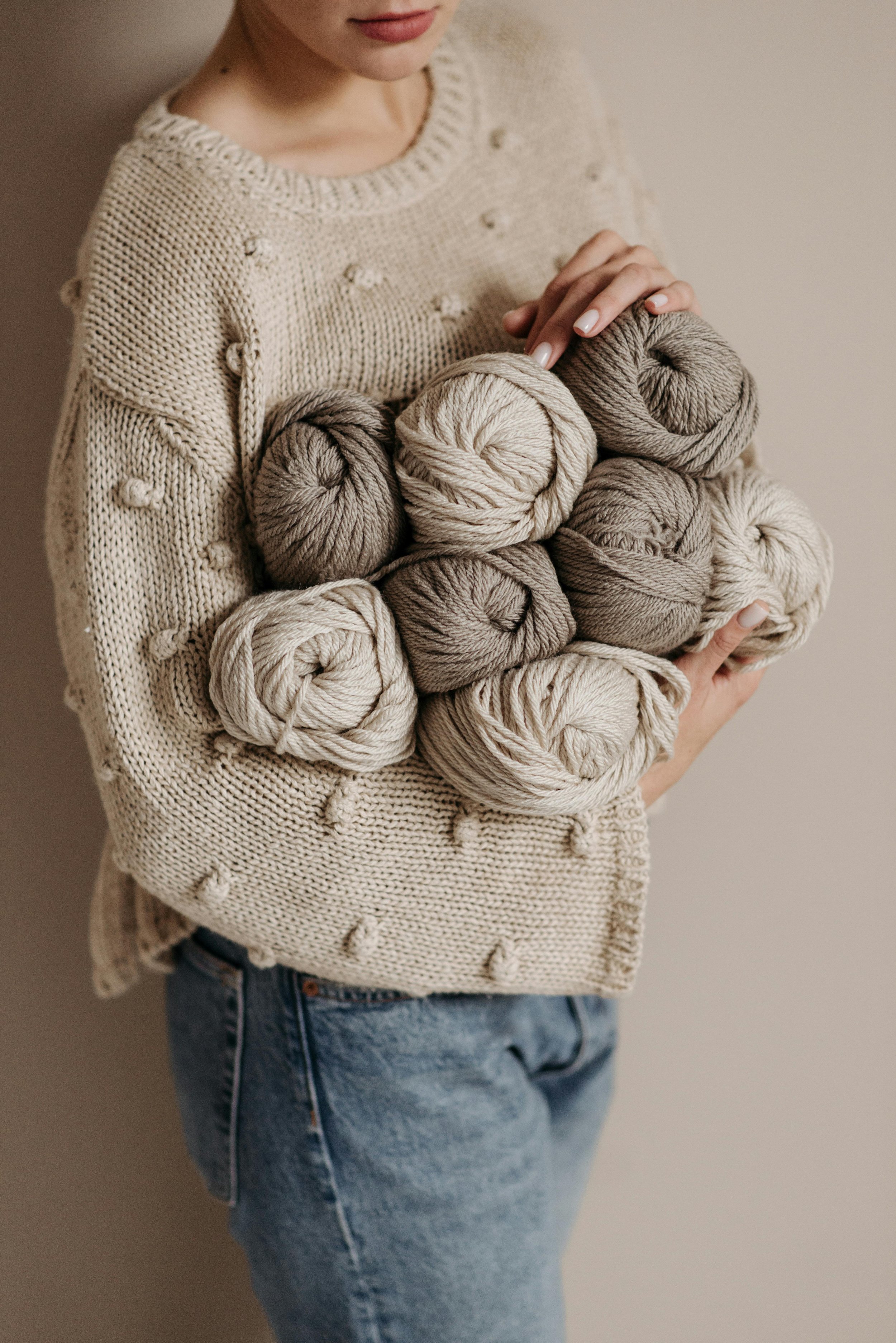 A woman wearing a beige knitted sweater holding ten skeins of yarn in neutral tones, standing against a plain background.