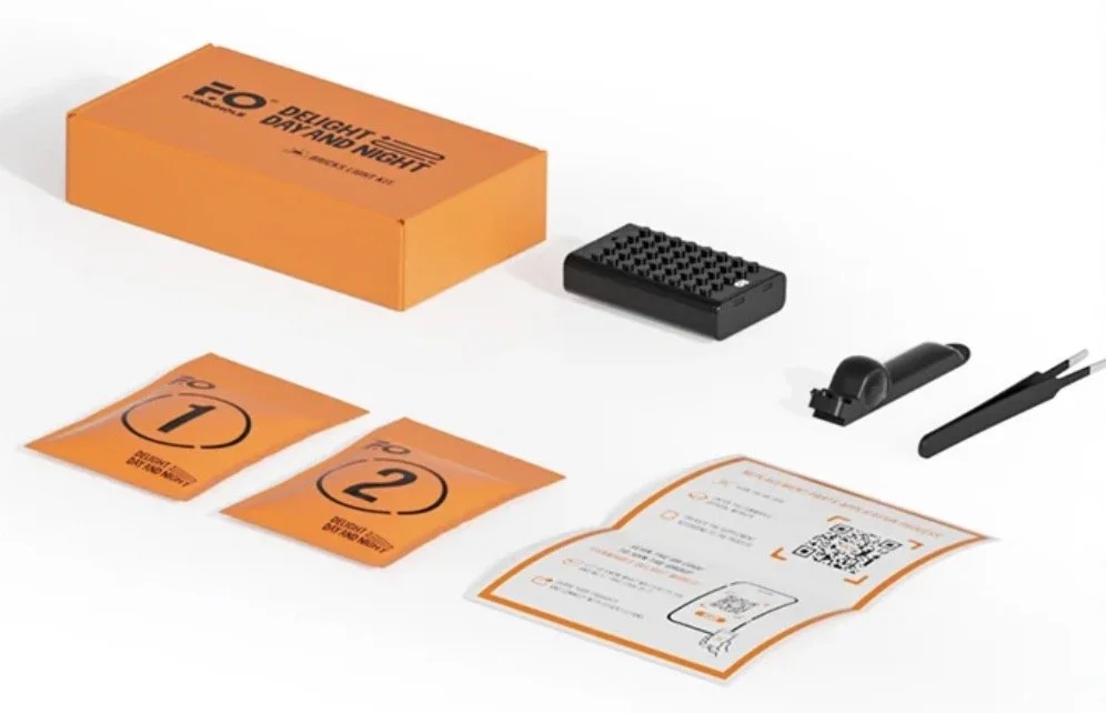 Kit de test de voyage avec boîte orange, instructions, cartes orange numérotées 1 et 2, un petit appareil noir, un stylo noir, et un rouleau de stickers ou étiquettes.