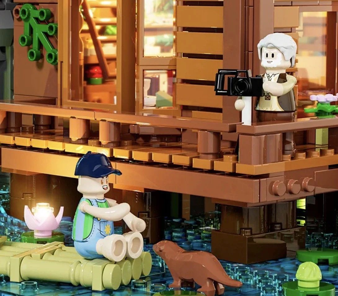 Une maison en briques LEGO avec deux personnages LEGO, un garçon assis près de l'eau avec un animal, et une femme avec un appareil photo à l'étage.