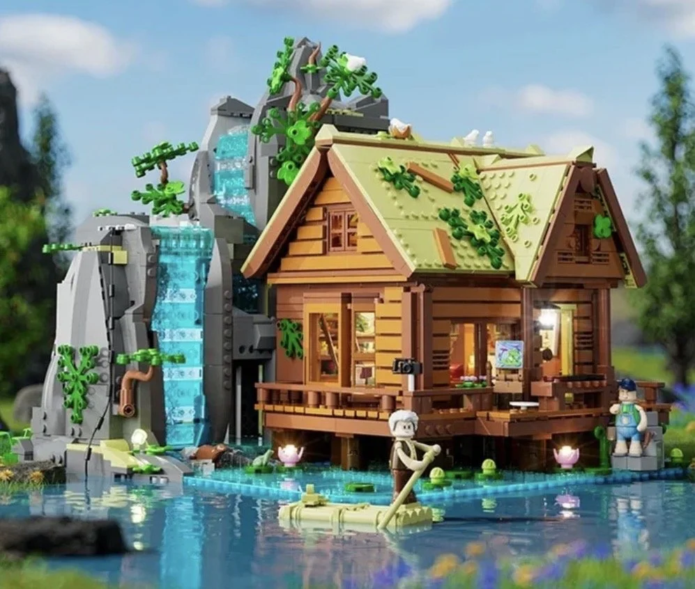 Modèle réduit en LEGO d'une maison en bois près d'une cascade et d'un étang, avec des personnages LEGO en train de faire du canoë et se détendre, dans un environnement naturel.