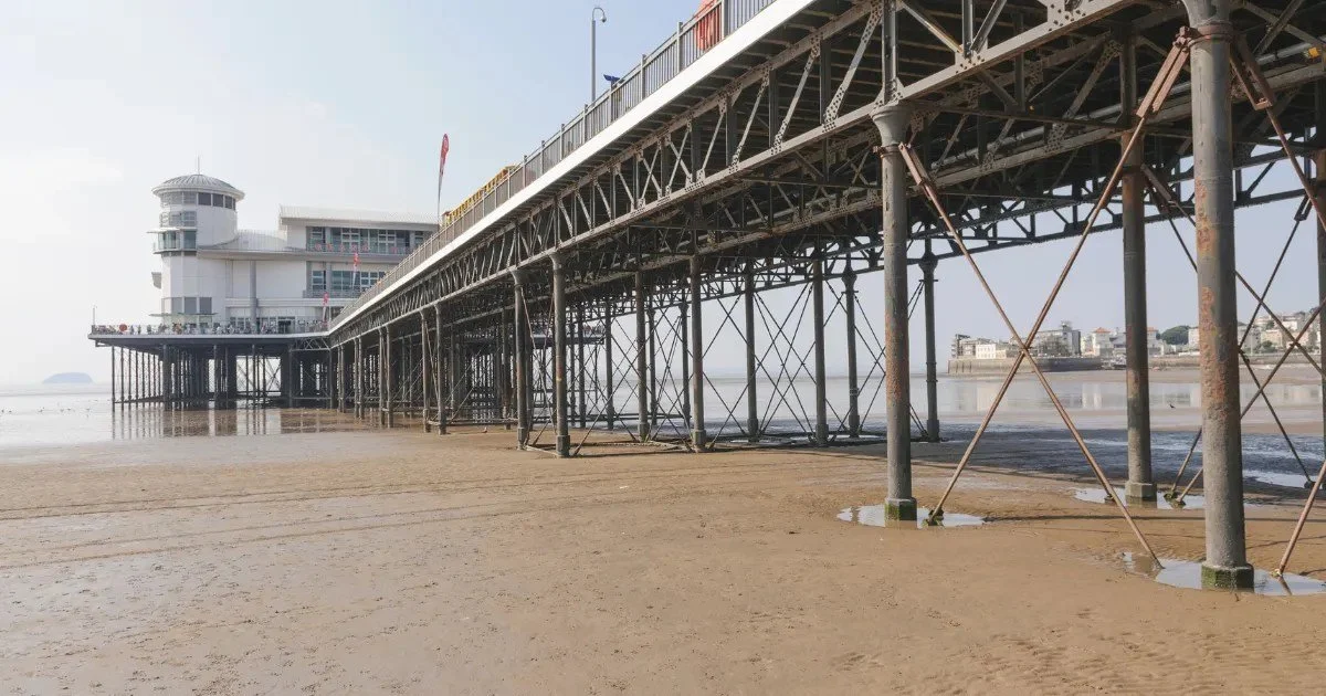 View of a pier extending over a sandy beach with a building at the end, under a clear sky. The pier's metal supports and structure are visible, and some buildings are seen in the distance.
