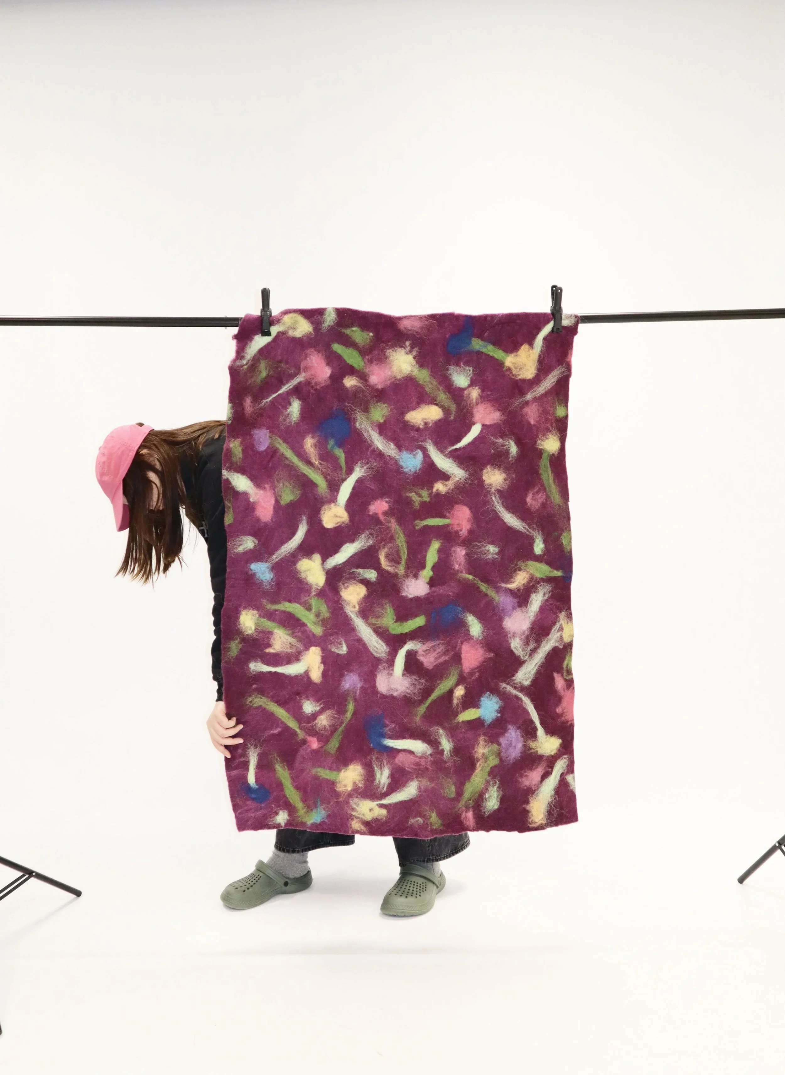 Child with brown hair wearing a pink hat and black clothes, holding a colorful felt fabric with a floral pattern, standing on a plain white background with photo studio equipment visible.