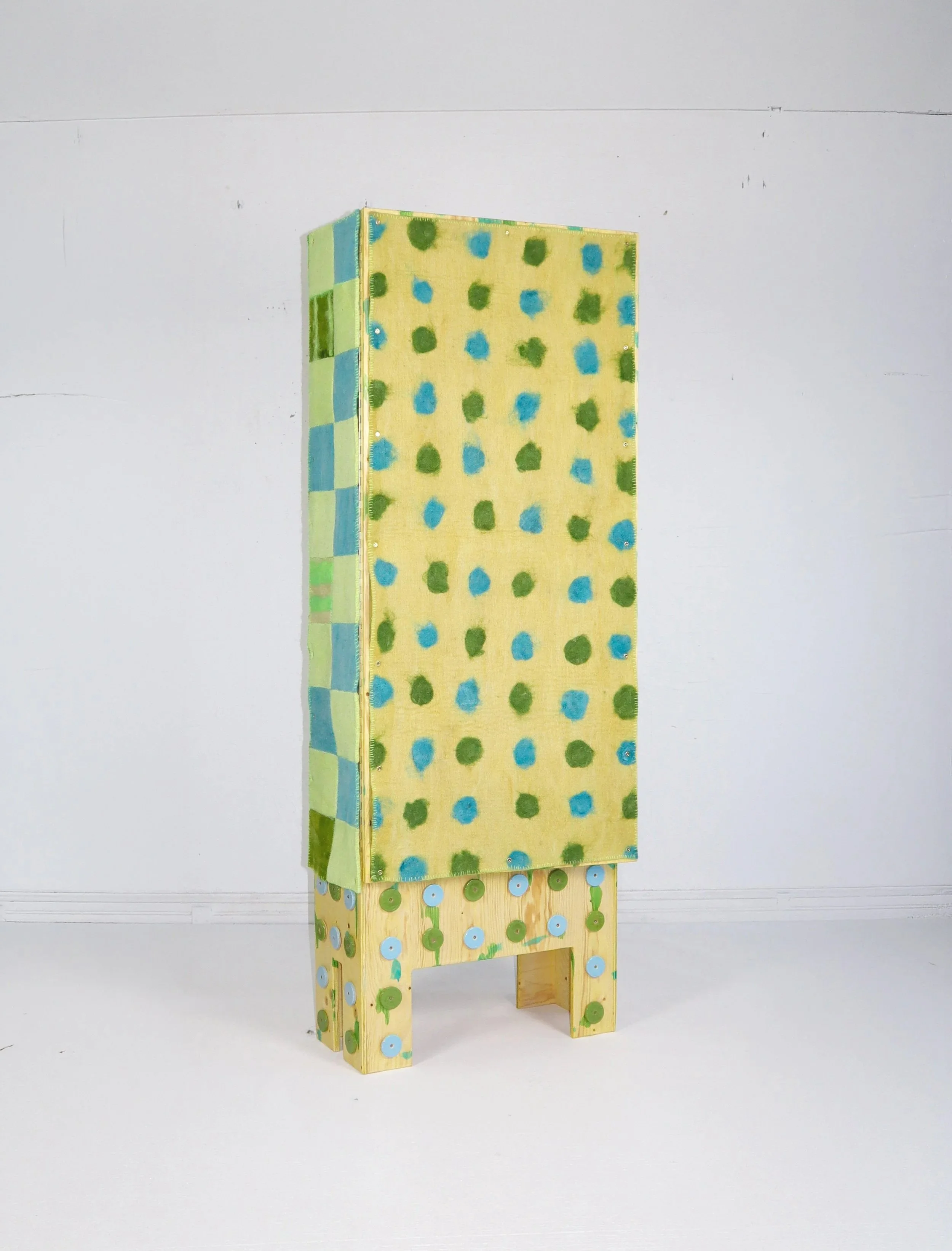 A colorful handmade box decorated with green and blue circle patterns and blue and green buttons, placed on a simple wooden stand against a plain white background.