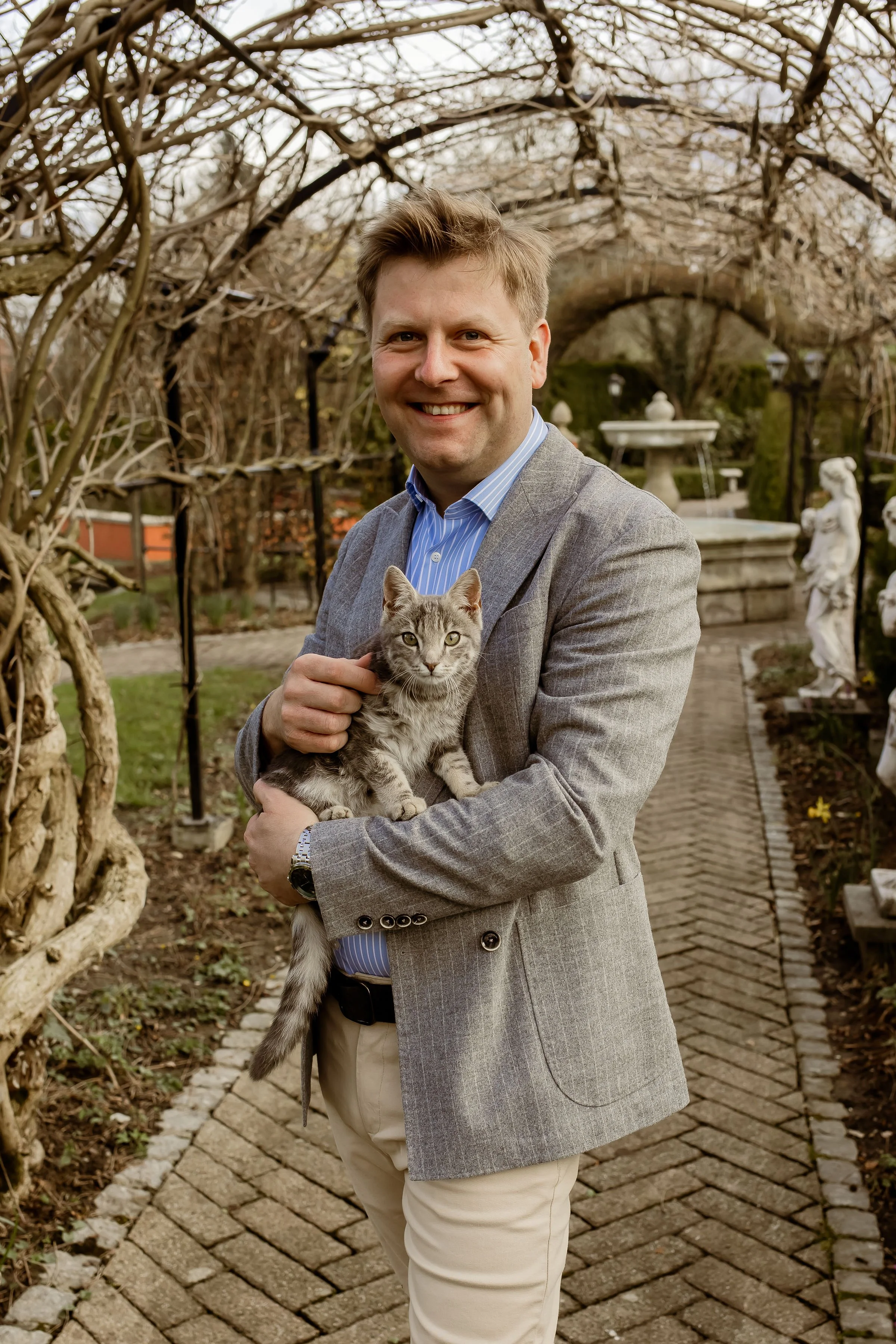 Ein lachender Mann in einem grauen Anzug hält eine kleine graue Katze in den Armen, im Hintergrund eine Gartenanlage mit Skulpturen, Brunnen und Bäumen.