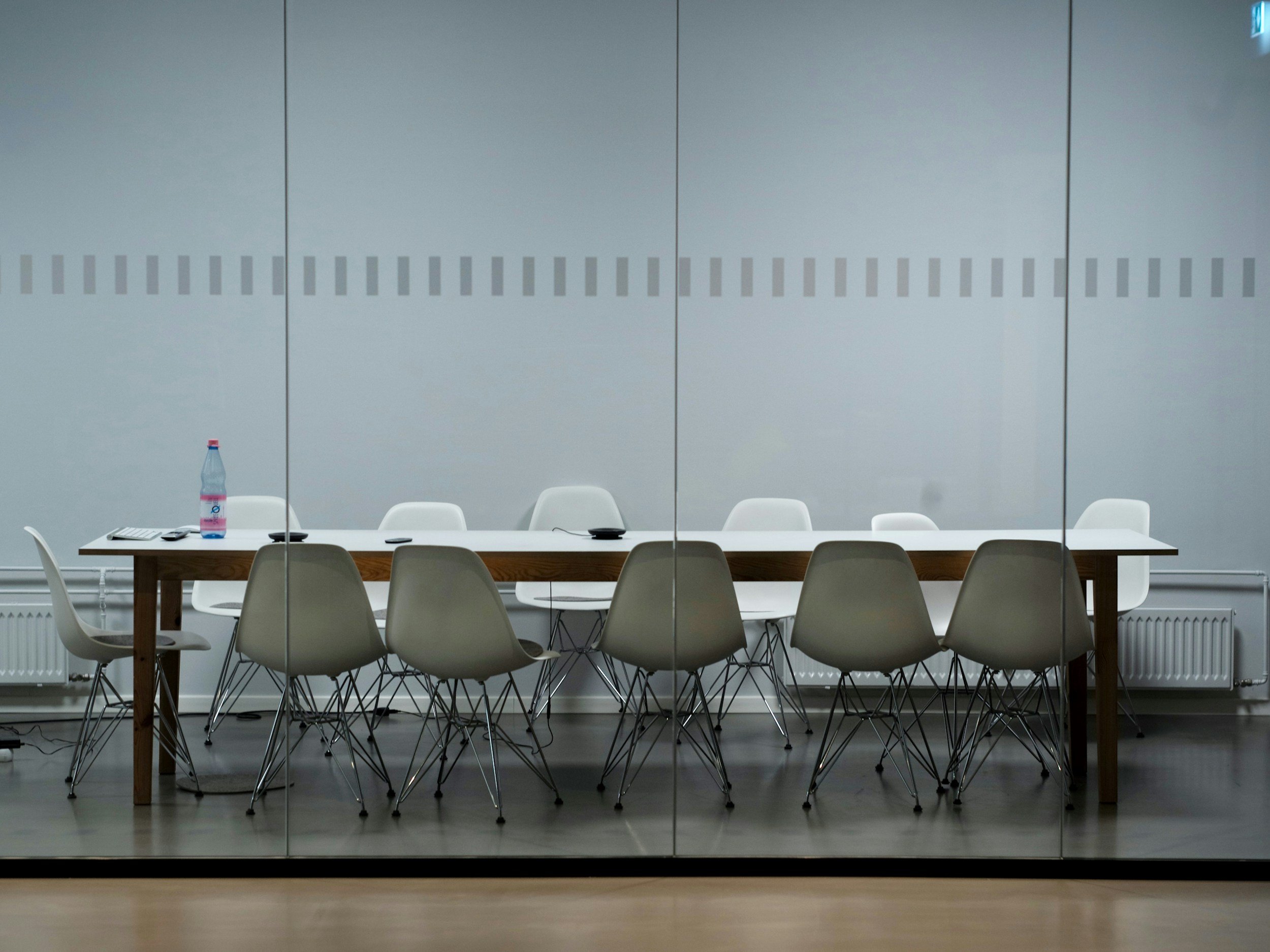 Empty conference room with a long wooden table, white chairs around it, one with a pink water bottle, and two black objects on the table. The room has a reflective glass wall and a gray wall with a dotted stripe pattern.