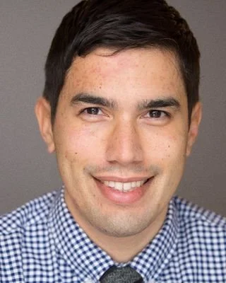 Close-up of a young man with dark hair, smiling, wearing a checkered shirt, against a plain background.