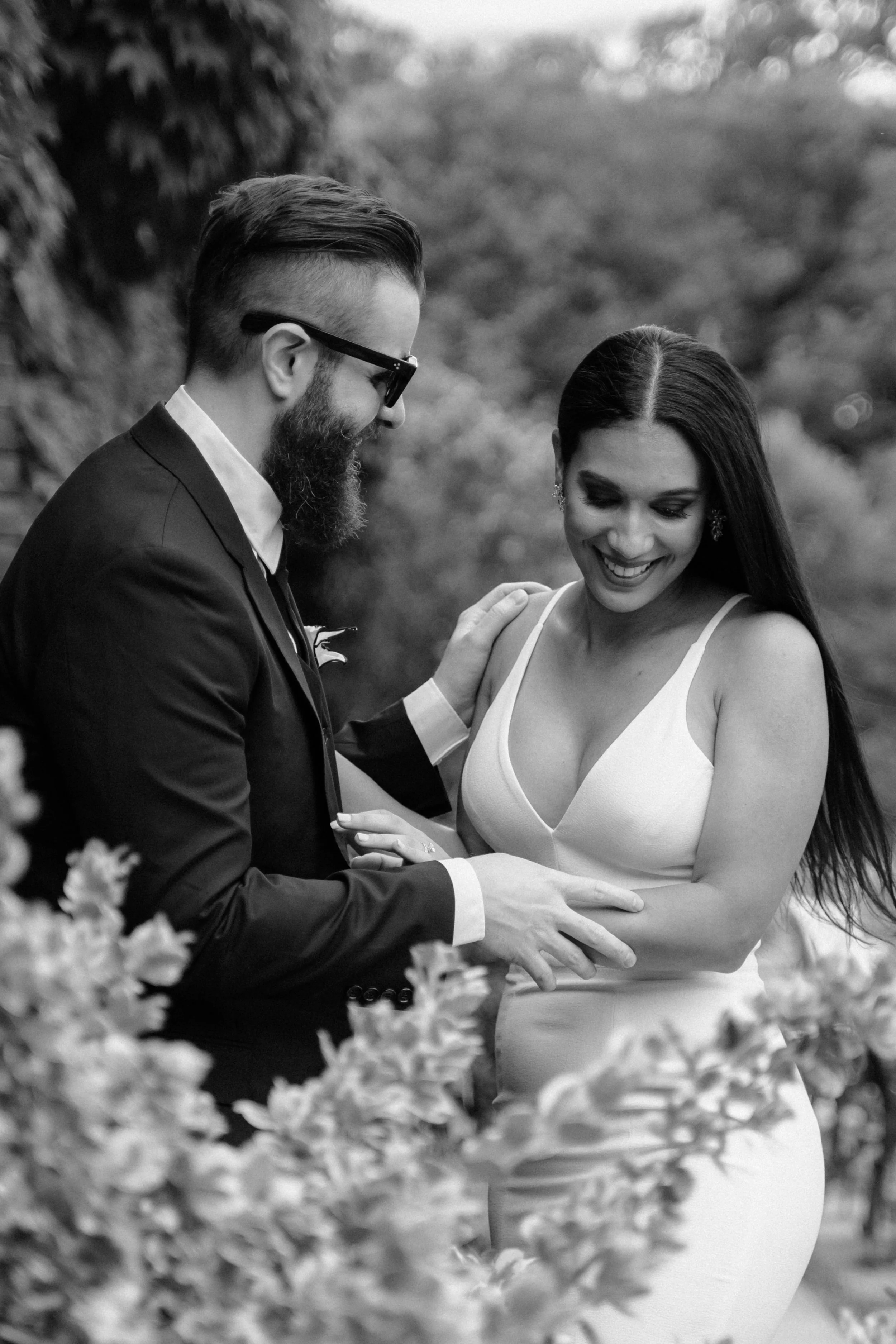 Una coppia sorridente, un uomo con barba, occhiali e abito scuro, e una donna con lunghi capelli scuri e vestito bianco, si abbracciano in un ambiente naturale in bianco e nero. Fotografa matrimonio Sardegna Costa Smeralda