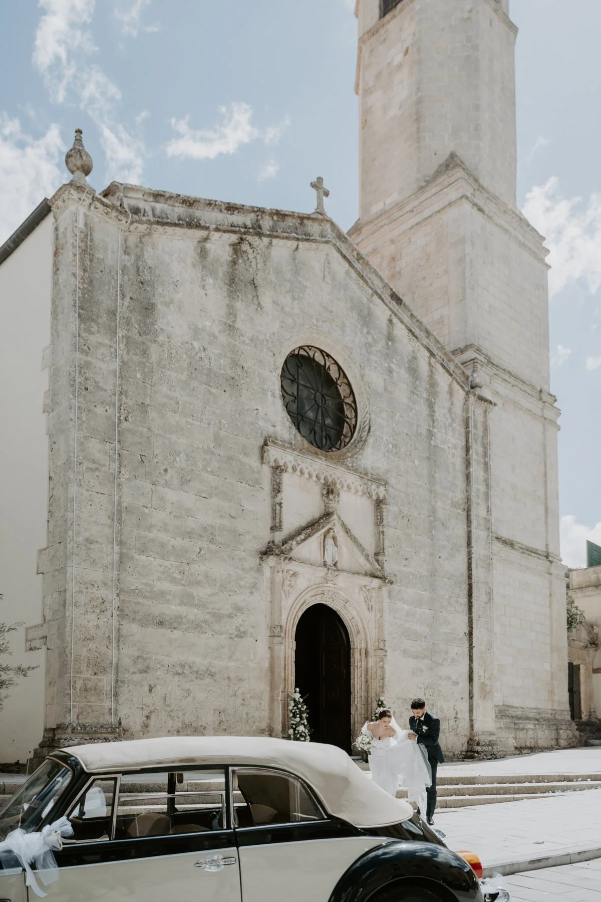 Una chiesa storica di pietra con una coppia di sposi che cammina davanti all'ingresso, accompagnata da una vettura d'epoca decorata con nastri di nozze Fotografa di matrimonio Sardegna