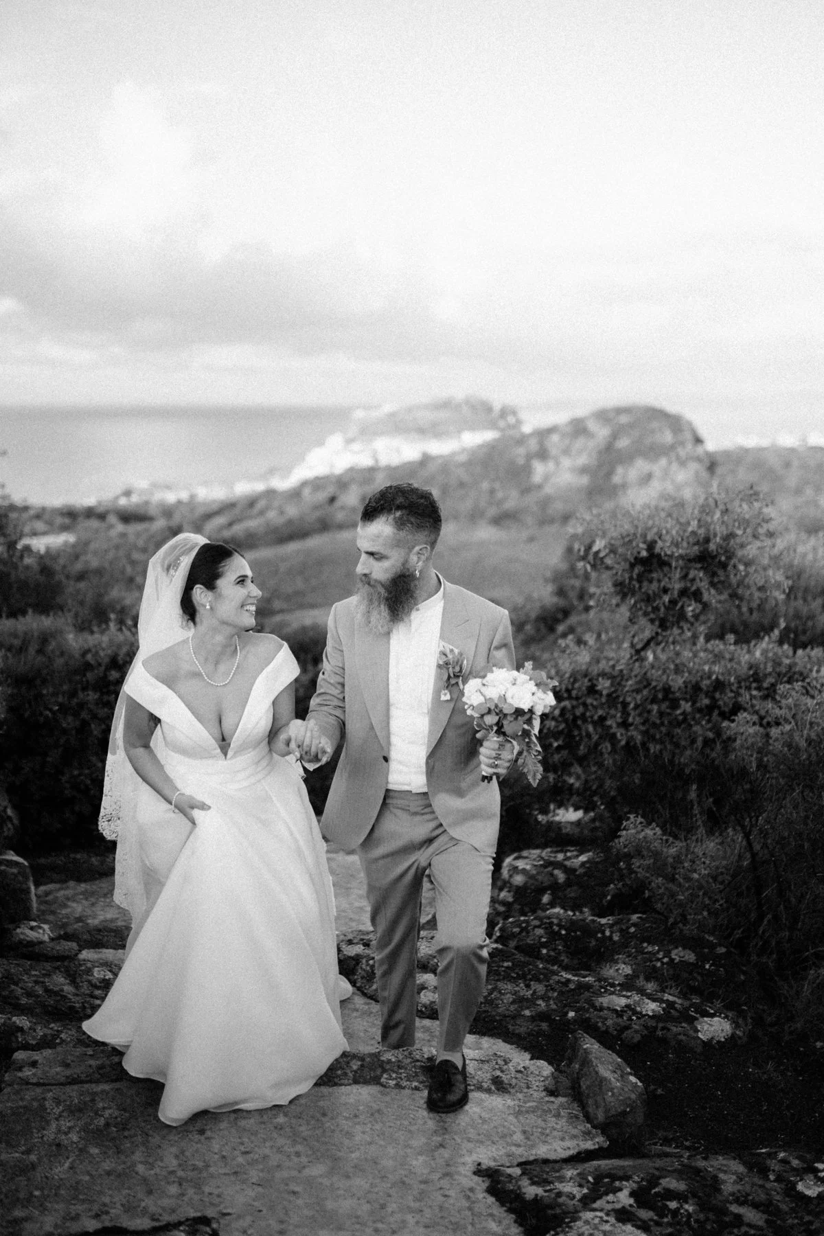 fotografa Daria Manuedda Una coppia di sposi cammina mano nella mano in un paesaggio naturale, sorridendo e guardandosi affettuosamente, durante il loro matrimonio.