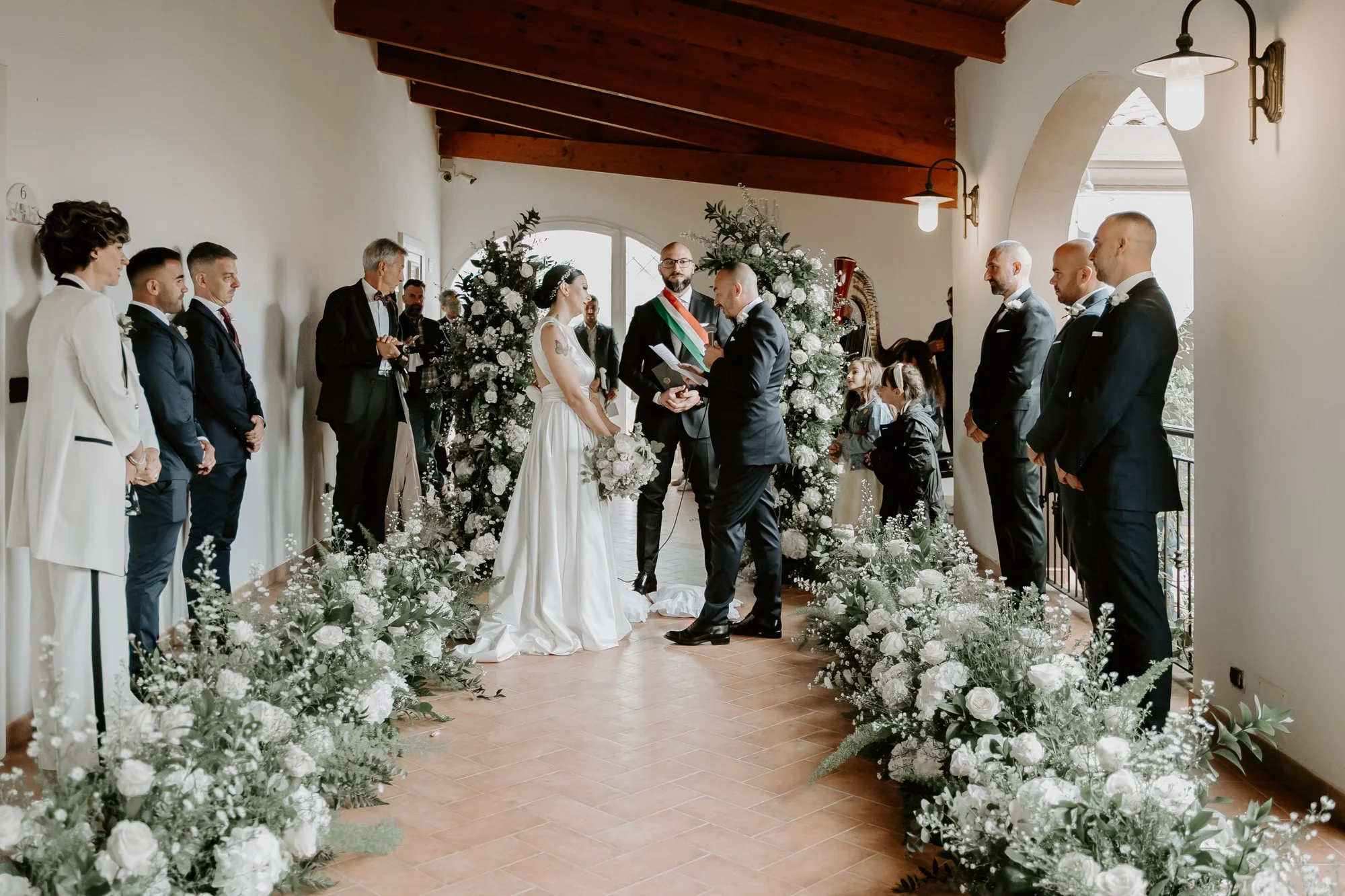 Cerimonia di matrimonio con sposi, testimoni e invitati in un ambiente decorato con fiori bianchi e piante, con pareti bianche e soffitto in legno.