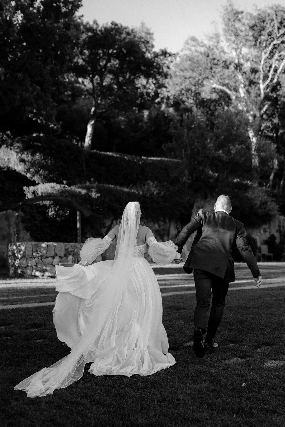 Una coppia di sposi, eguagliata con abiti da matrimonio, corre mano nella mano su un prato; lo sfondo mostra alberi e vegetazione in un ambiente naturale.