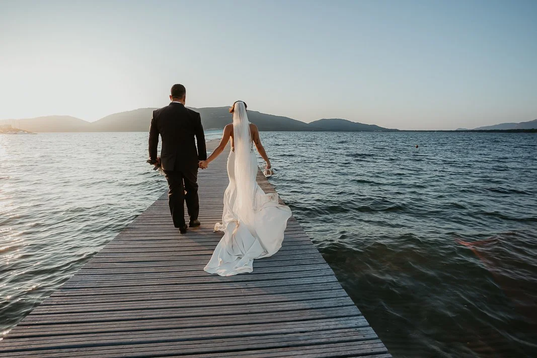 Una coppia di sposi cammina su un pontile sul lago al tramonto, con montagne sullo sfondo.
