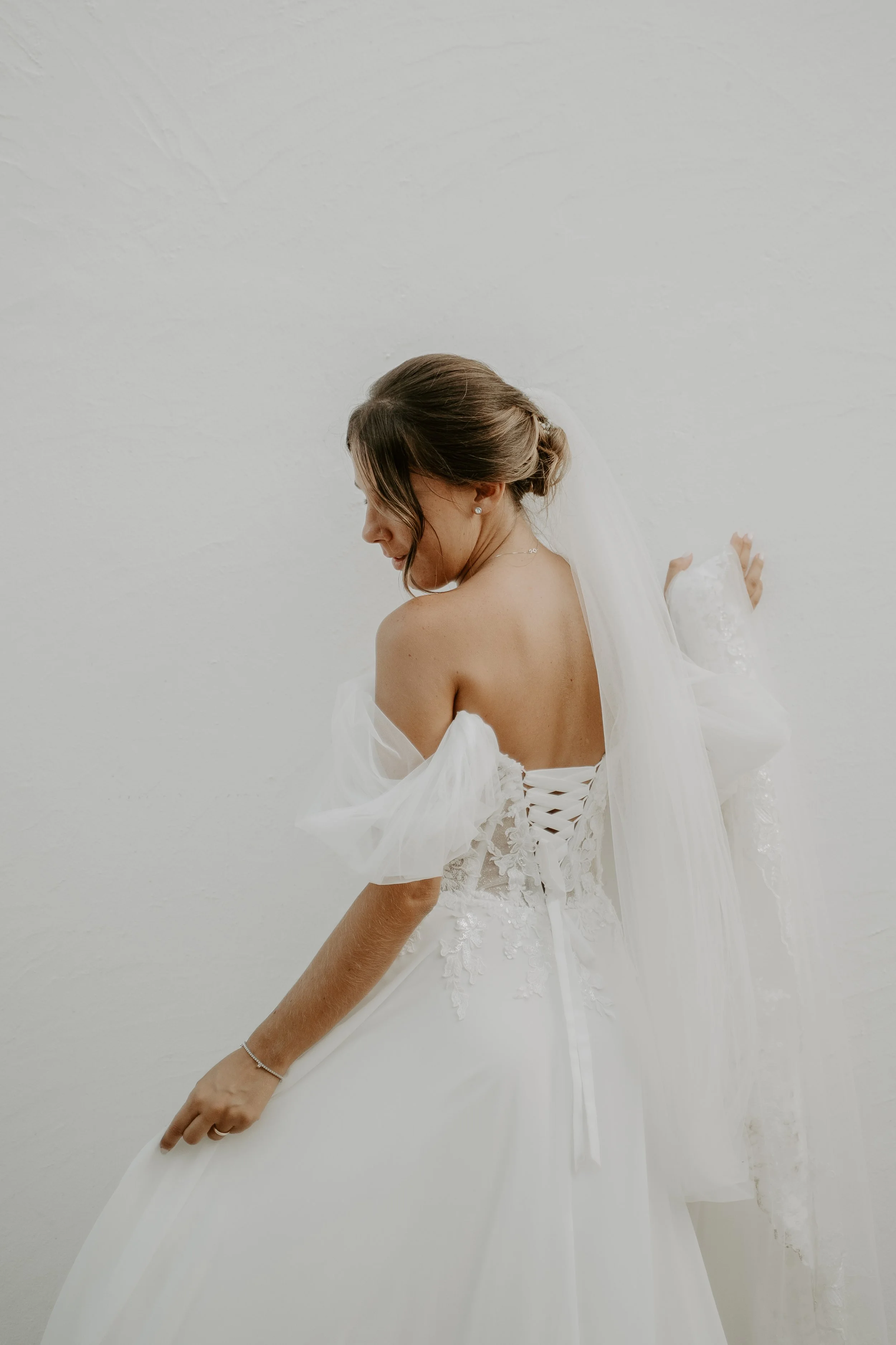 Una donna in abito da sposa bianco con dettagli di pizzo che sorride e guarda verso il basso, posando davanti a una parete bianca.