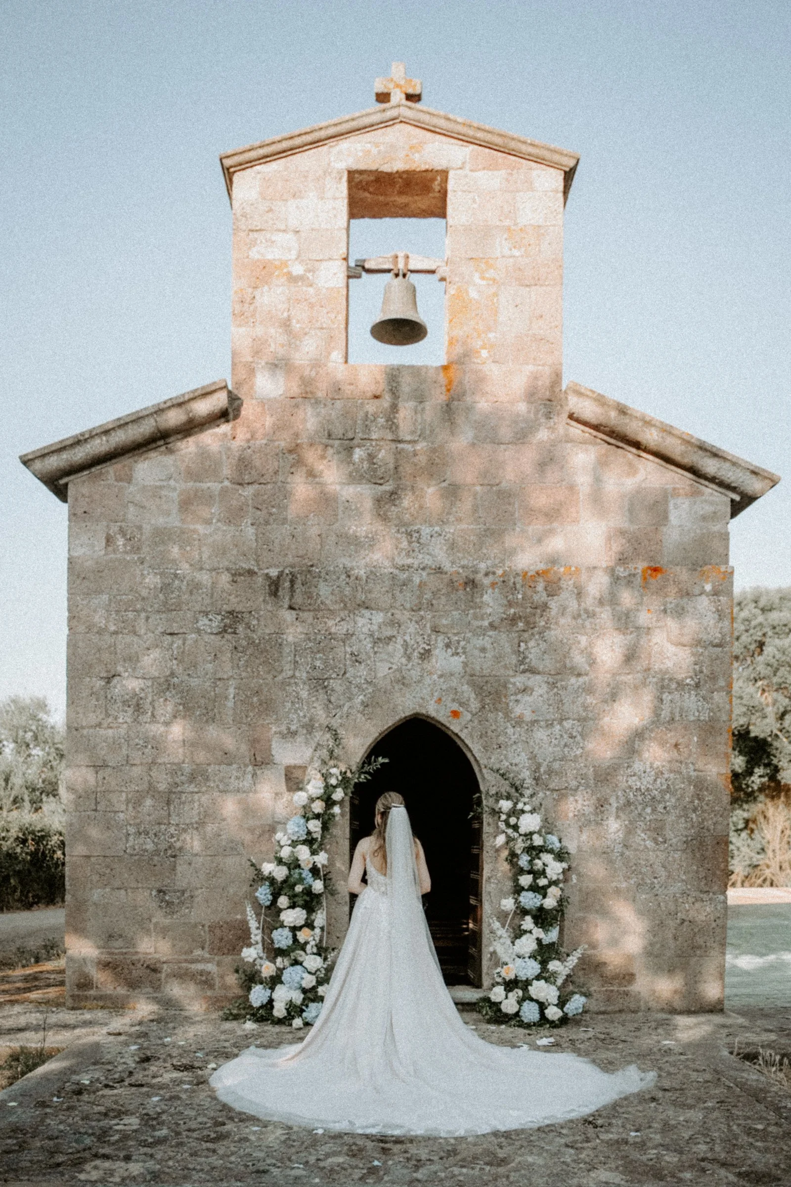 Una sposa in abito bianco con lungho velo si trova davanti a una chiesa di pietra decorata con fiori. È vista da dietro mentre si avvicina all'ingresso.