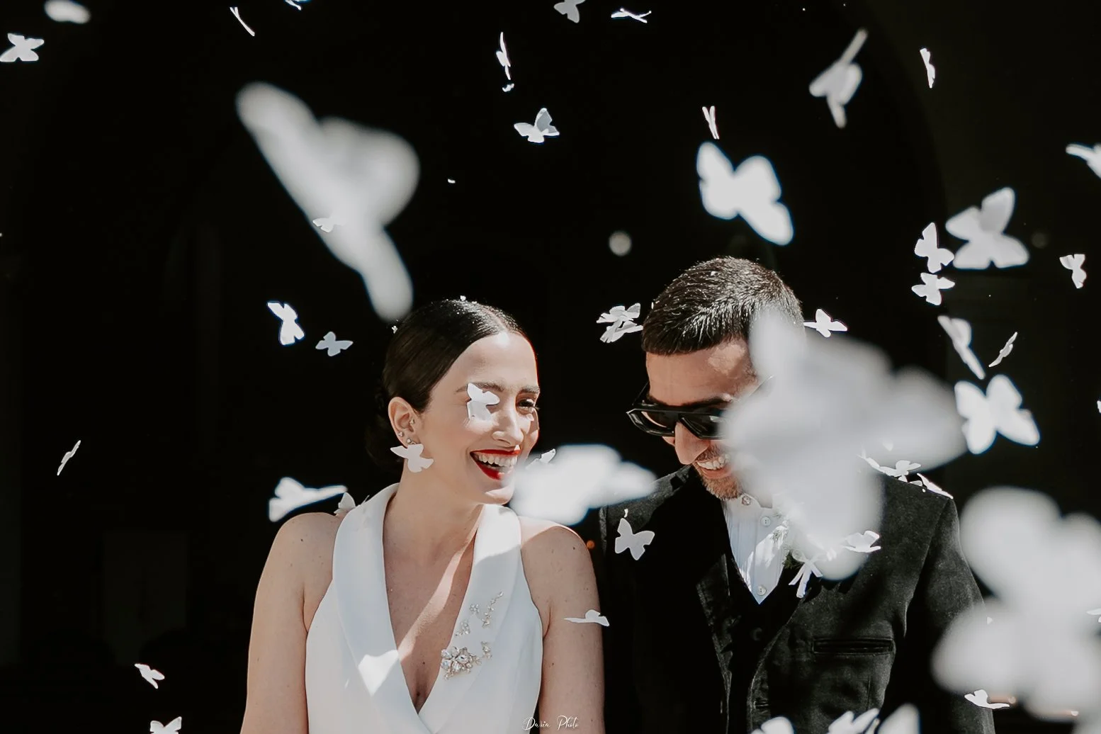 Una coppia sorridente, una donna in abito bianco e un uomo in giacca nera, circondati da farfalle di carta bianche, in un'atmosfera gioiosa. fotografa matrimonio Sardegna Castelsardo