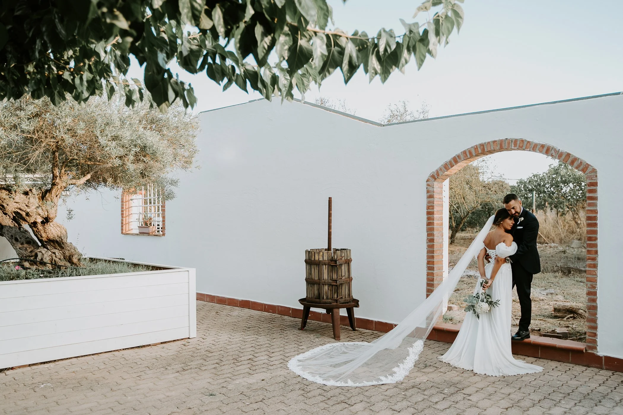 Una coppia ne areta celebra un matrimonio all'aperto, con la sposa che indossa un abito bianco con un lungo velo e il bouquet, mentre il sposo, vestito di nero, la tiene stretta. Sono sotto un arco di mattoni, circondati da un paesaggio naturale con alberi e un cielo sereno.