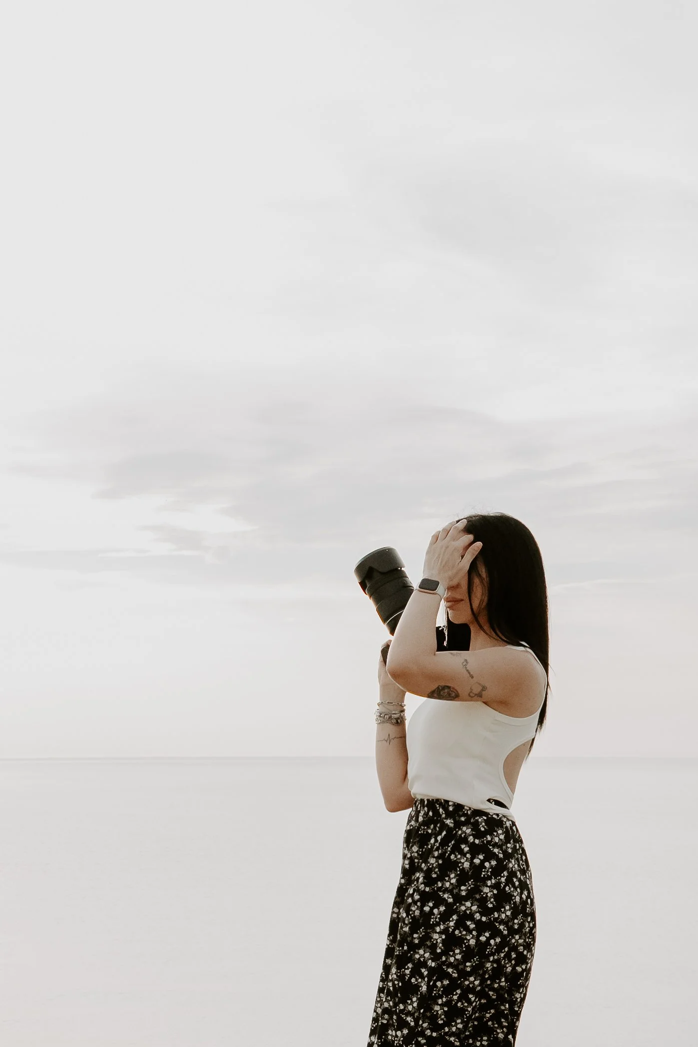 Una donna con tatuaggi, che indossa una maglietta bianca e una gonna lunga a fiori, tiene una fotocamera al collo e si sistema i capelli, davanti a un cielo nuvoloso.