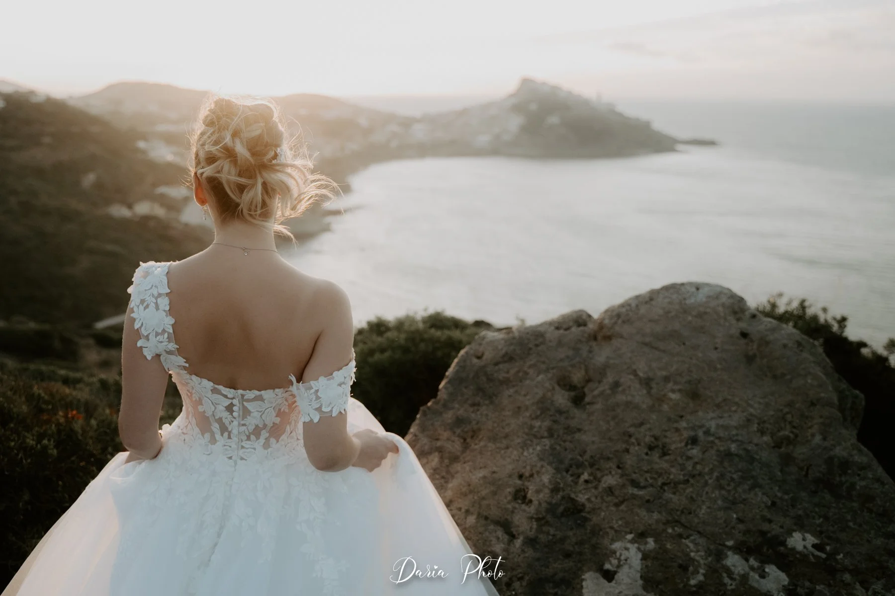 Una donna in un abito da sposa bianco guarda il mare al tramonto, con le onde e il paesaggio costiero sullo sfondo.