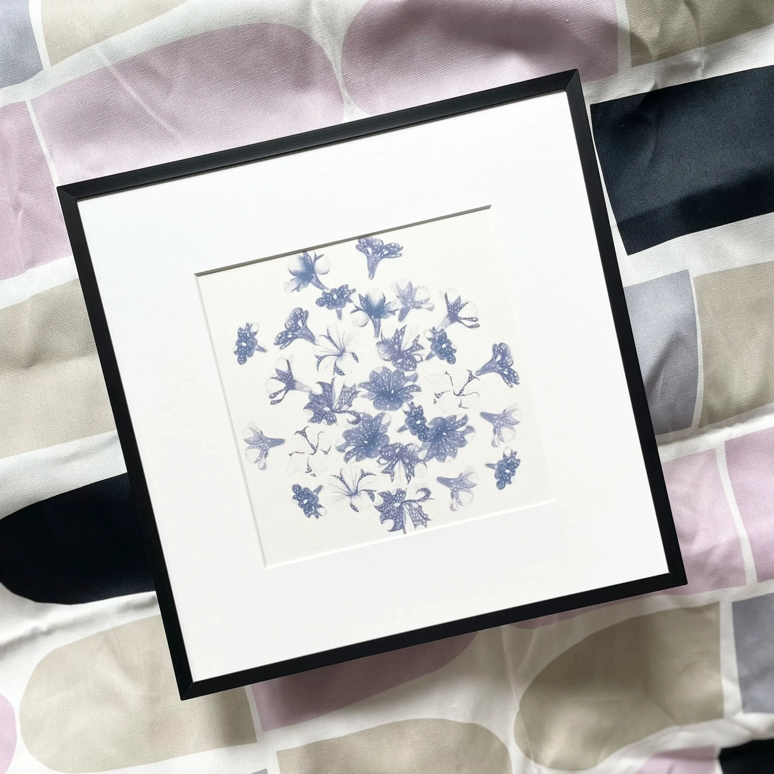 Framed artwork of blue flowers and butterflies on a white background, resting on a patterned fabric surface.