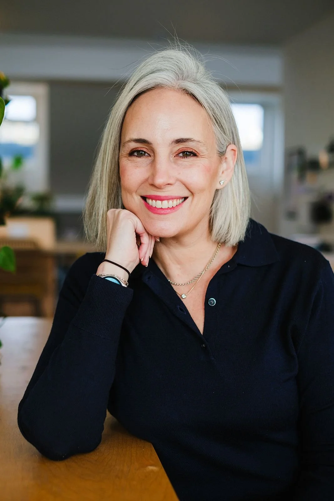 A woman with shoulder-length gray hair smiling, resting her chin on her hand, wearing a black shirt with a necklace, sitting at a wooden table indoors with windows in the background.