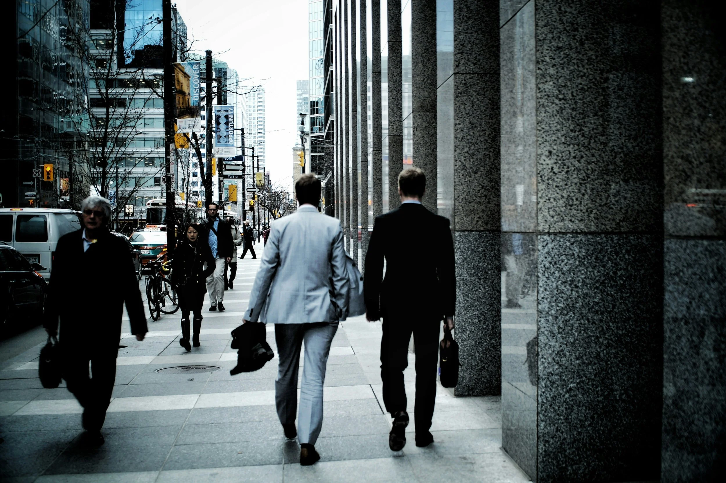 Two businessmen wearing suits walk together on a city sidewalk, surrounded by other pedestrians, tall buildings, and urban street activity.
