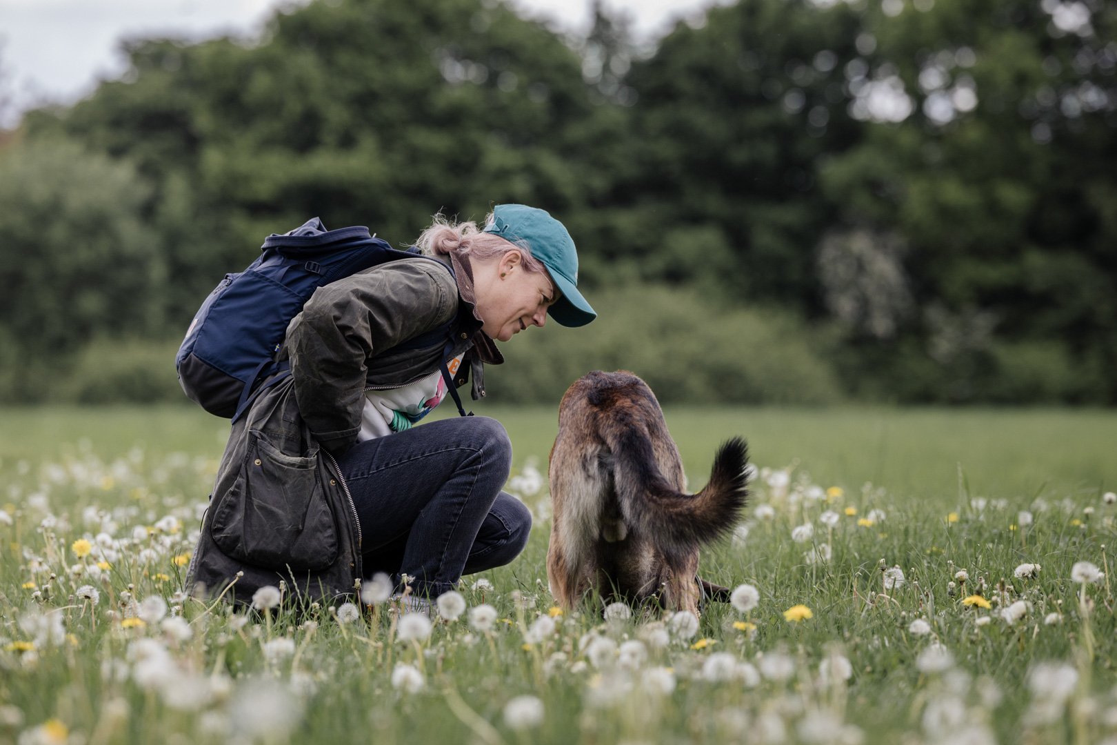 Eine Frau mit Rucksack kniet in einer Wiese voller Blumen und schaut aufmerksam auf ein Tier, das auf dem Boden sitzt, im Freien mit Bäumen im Hintergrund.