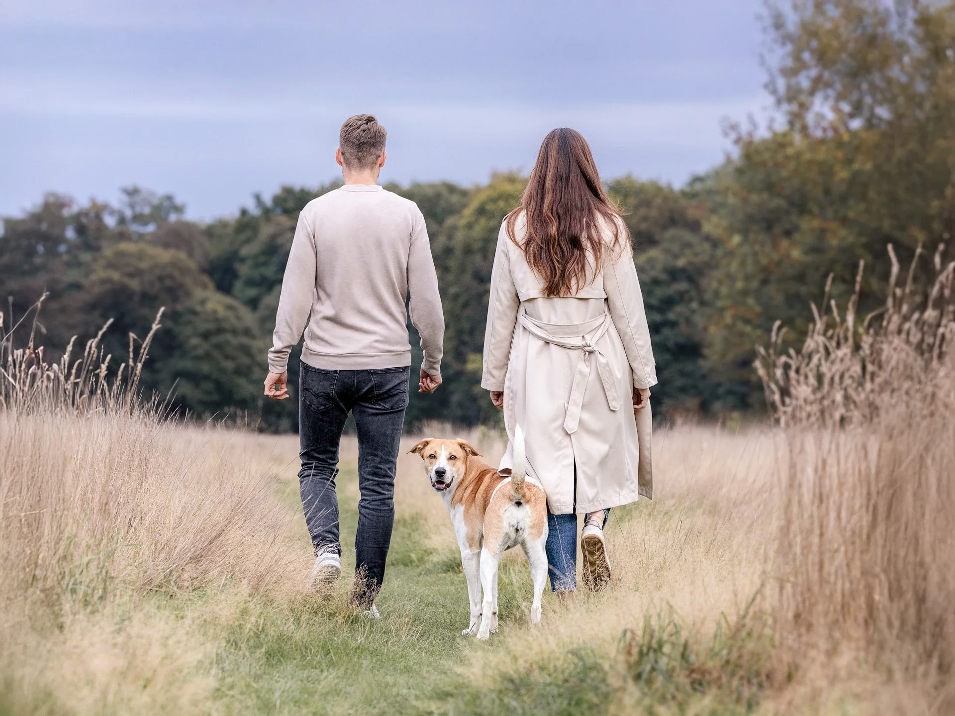 Zwei Menschen und ein Hund, die zusammen in einer Wiese spazieren gehen, mit Bäumen im Hintergrund.