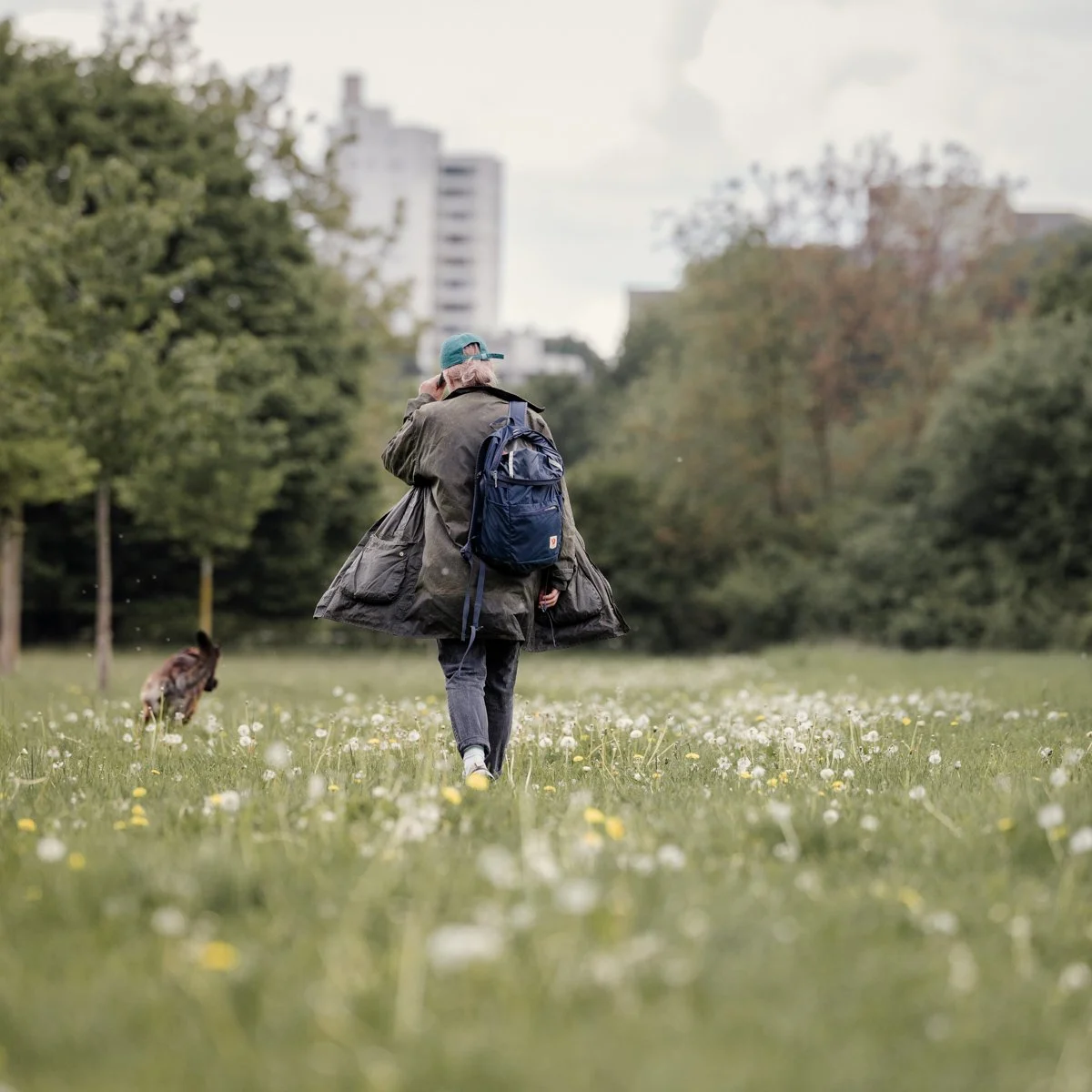 Eine Person mit Rucksack läuft durch ein Wiesenfeld mit Gänseblümchen, während ein Hund neben ihr läuft. Im Hintergrund sind Bäume und hohe Gebäude sichtbar.