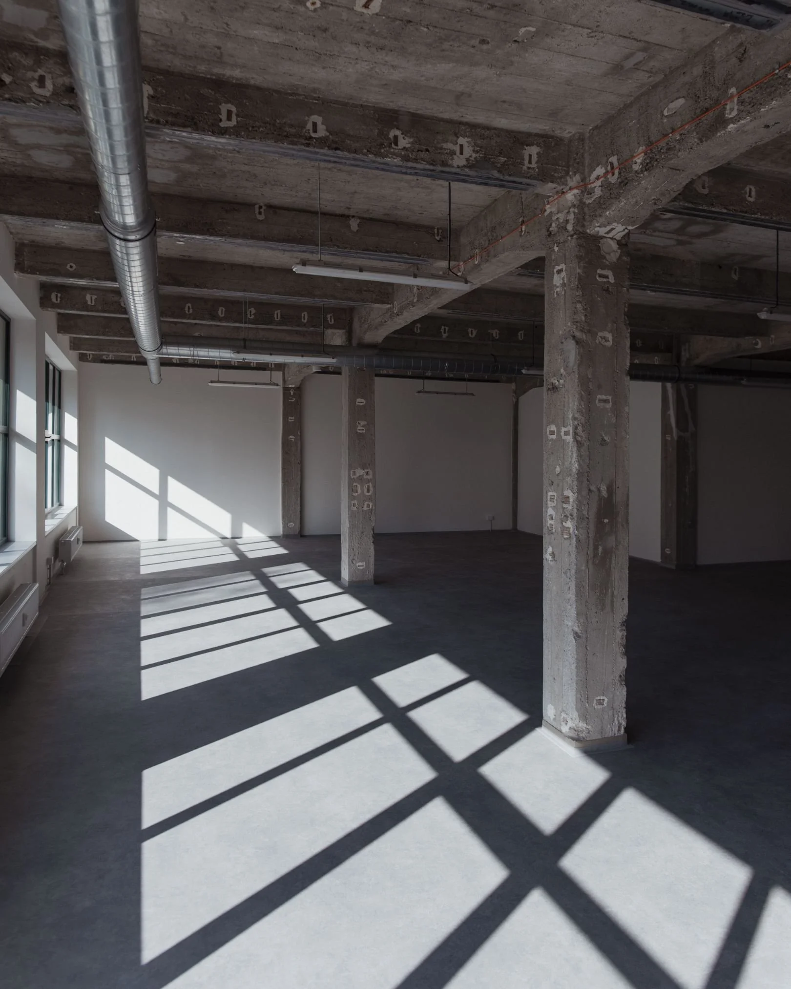 Empty industrial-style room with concrete pillars, exposed ductwork, and large windows casting shadows on the floor.