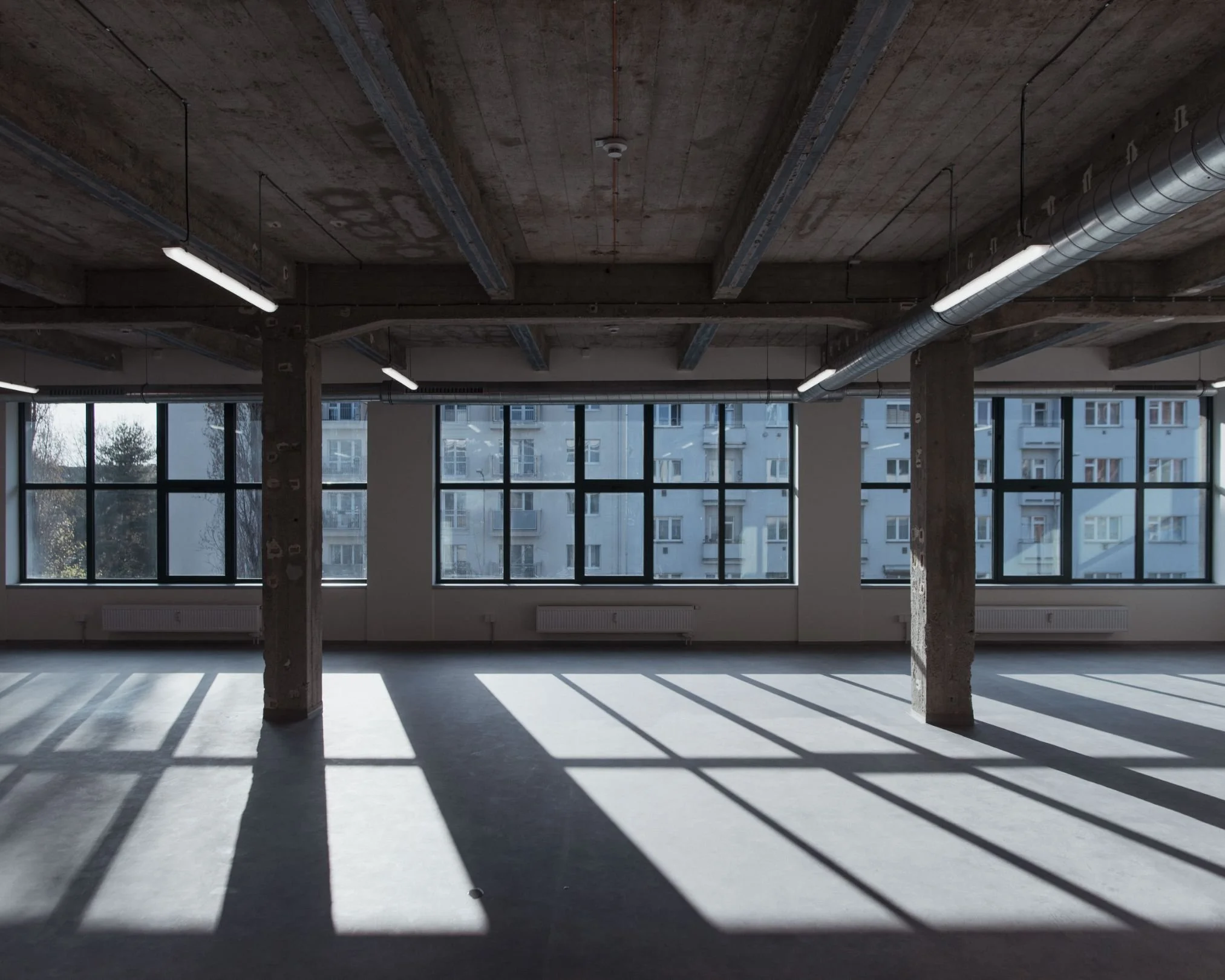 Empty room with large windows casting shadows on the floor, exposed ceiling with beams and ductwork.