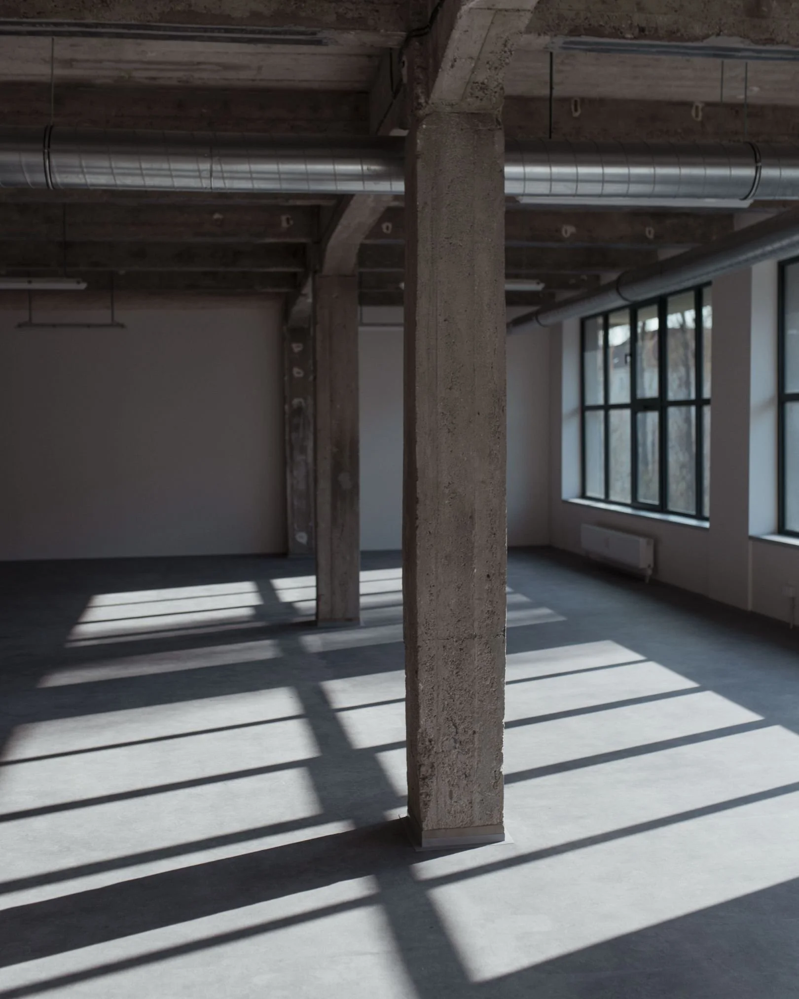 Empty industrial-style room with concrete pillars, large windows, and shadows on the floor.