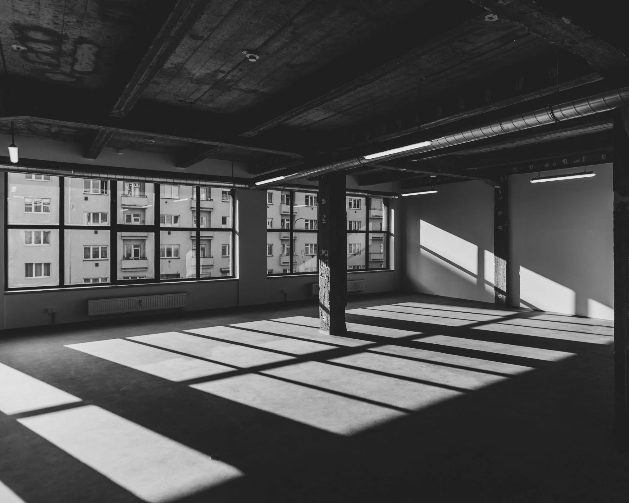 An empty, unfinished room with large windows casting shadows on the floor, exposed ceiling with beams and pipes.
