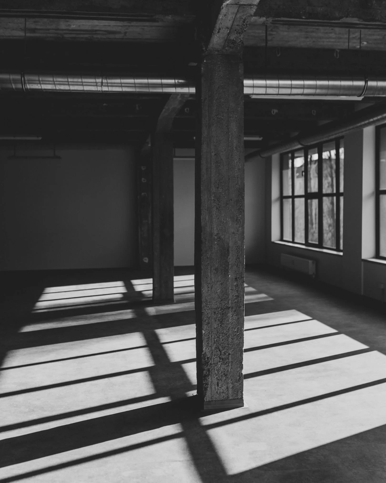 Empty room with large windows, concrete pillars, and shadows cast by window frames on the floor in black and white.