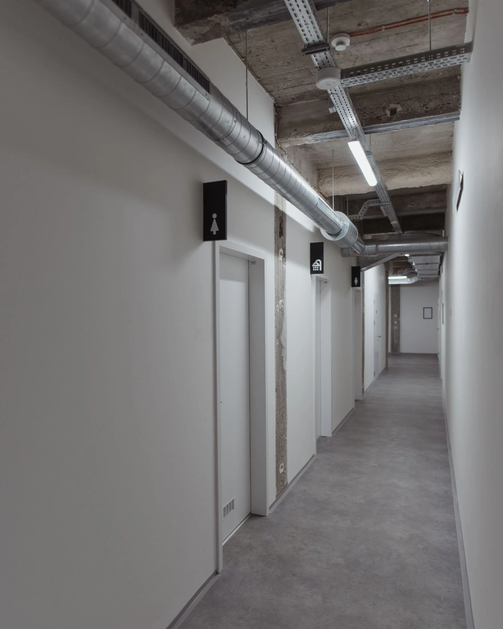 Empty indoor corridor with exposed ceiling pipes and signs for restrooms, stairs, and elevator.