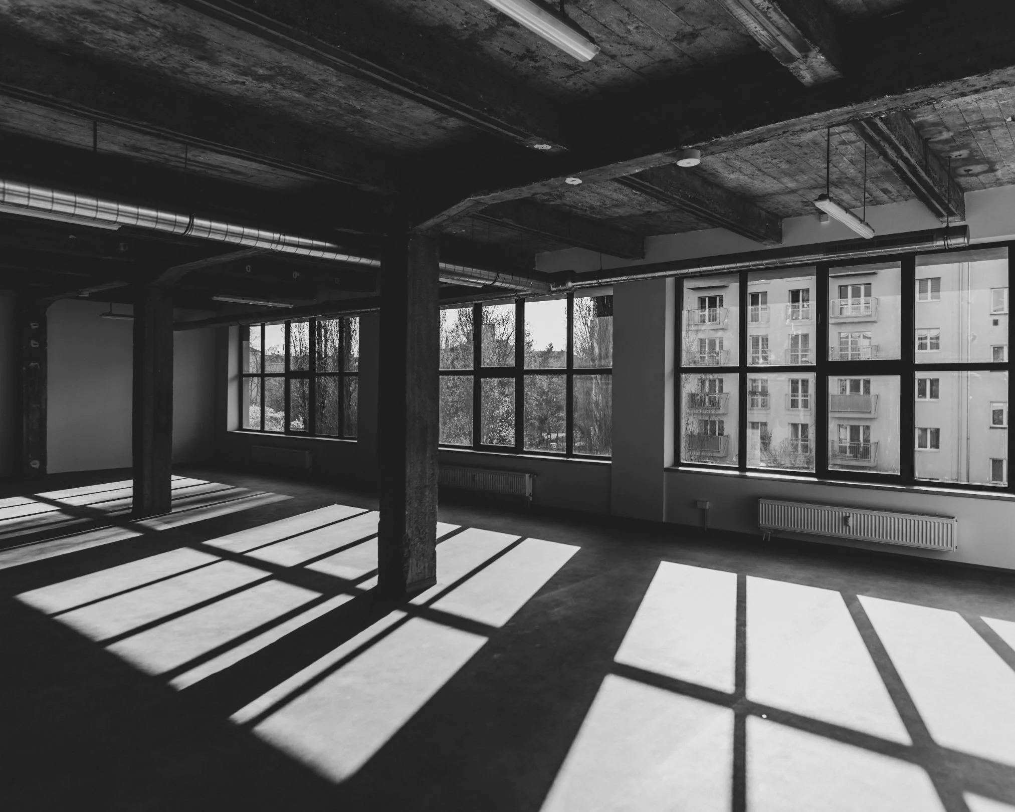 Empty industrial-style room with large windows and concrete ceiling, casting shadows on the floor.