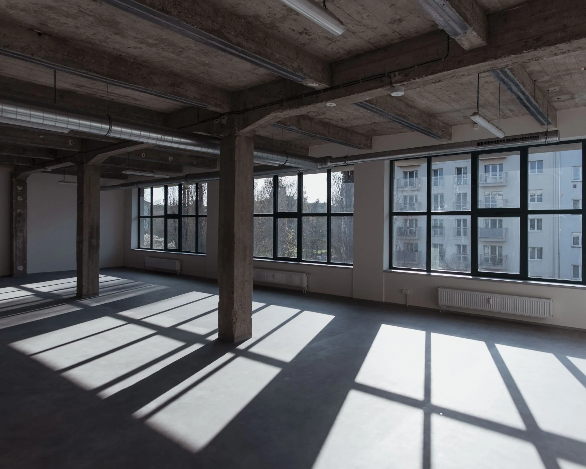 Empty loft with large windows casting shadows on the concrete floor, exposed ceiling with ducts, and structural columns.