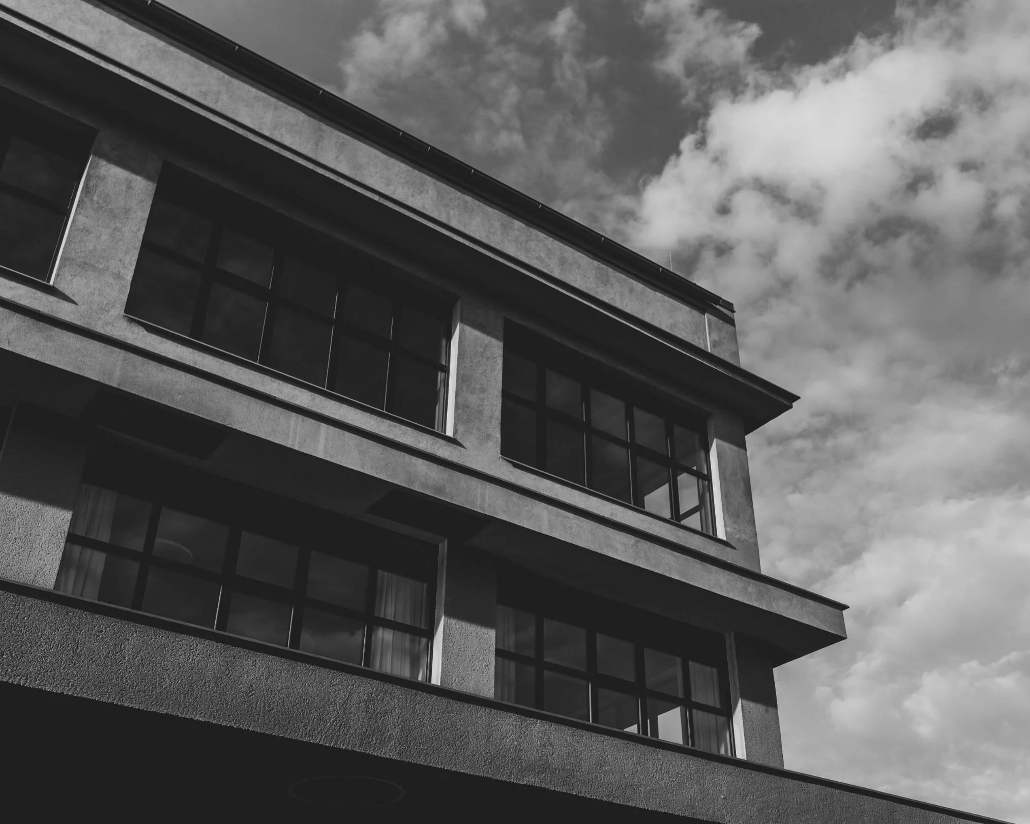 A black and white photo of a modern multi-story building with large glass windows and flat balconies, under a partly cloudy sky.