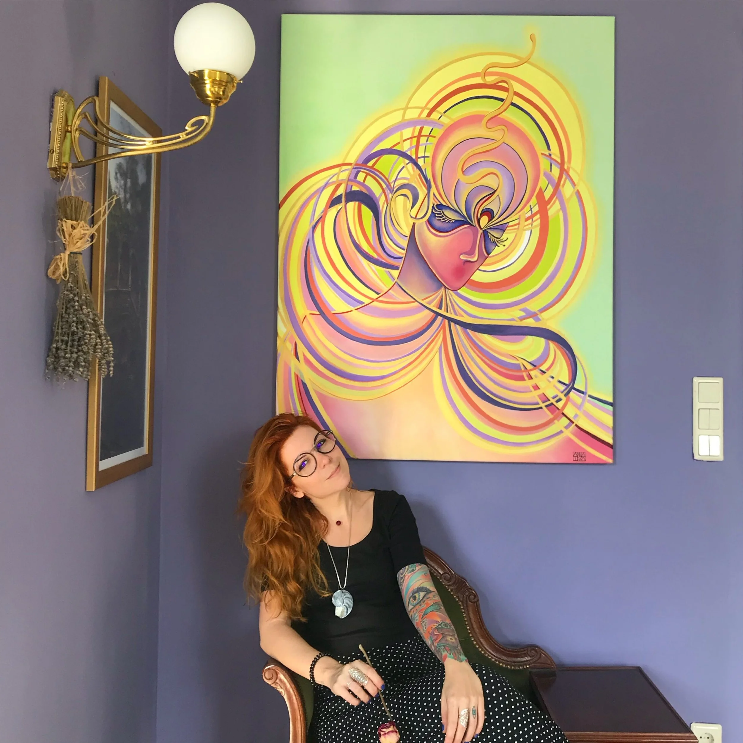 A woman with long red hair, glasses, and tattoos on her arm, sitting on a vintage wooden chair in front of a vibrant abstract painting of a woman's face with swirling lines and bright colors, in a room with purple walls and gold wall fixtures.