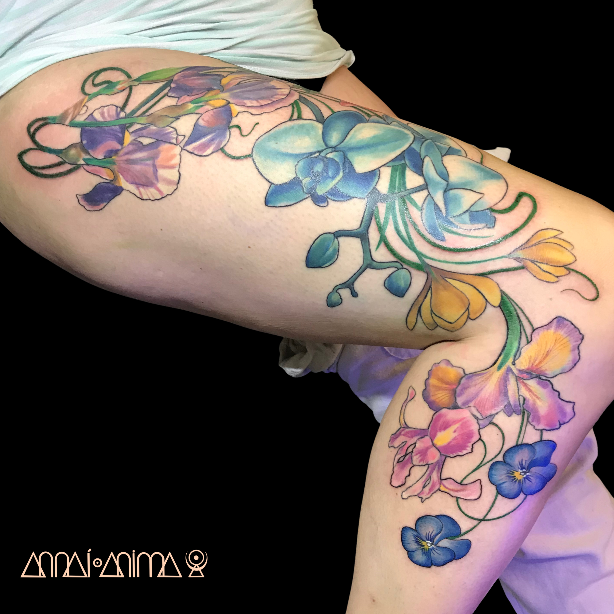 A colorful floral tattoo featuring purple, blue, yellow, and pink flowers on a person's arm and leg, with intricate details and shading.