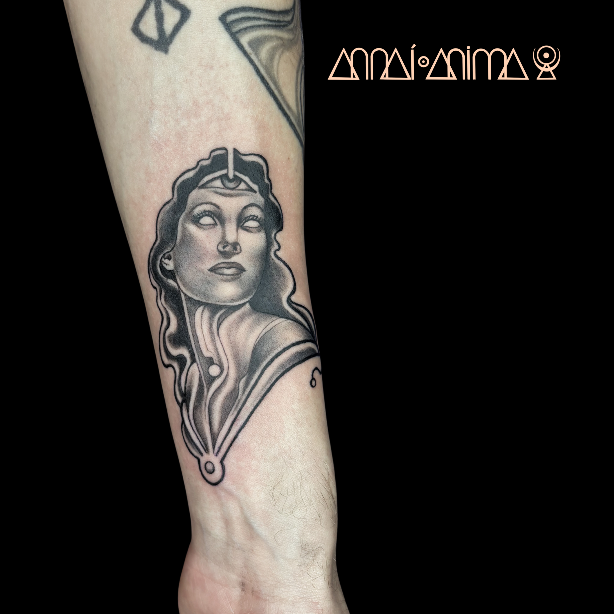 Black and gray tattoo of a woman with three eyes, long wavy hair, and abstract decorative elements.