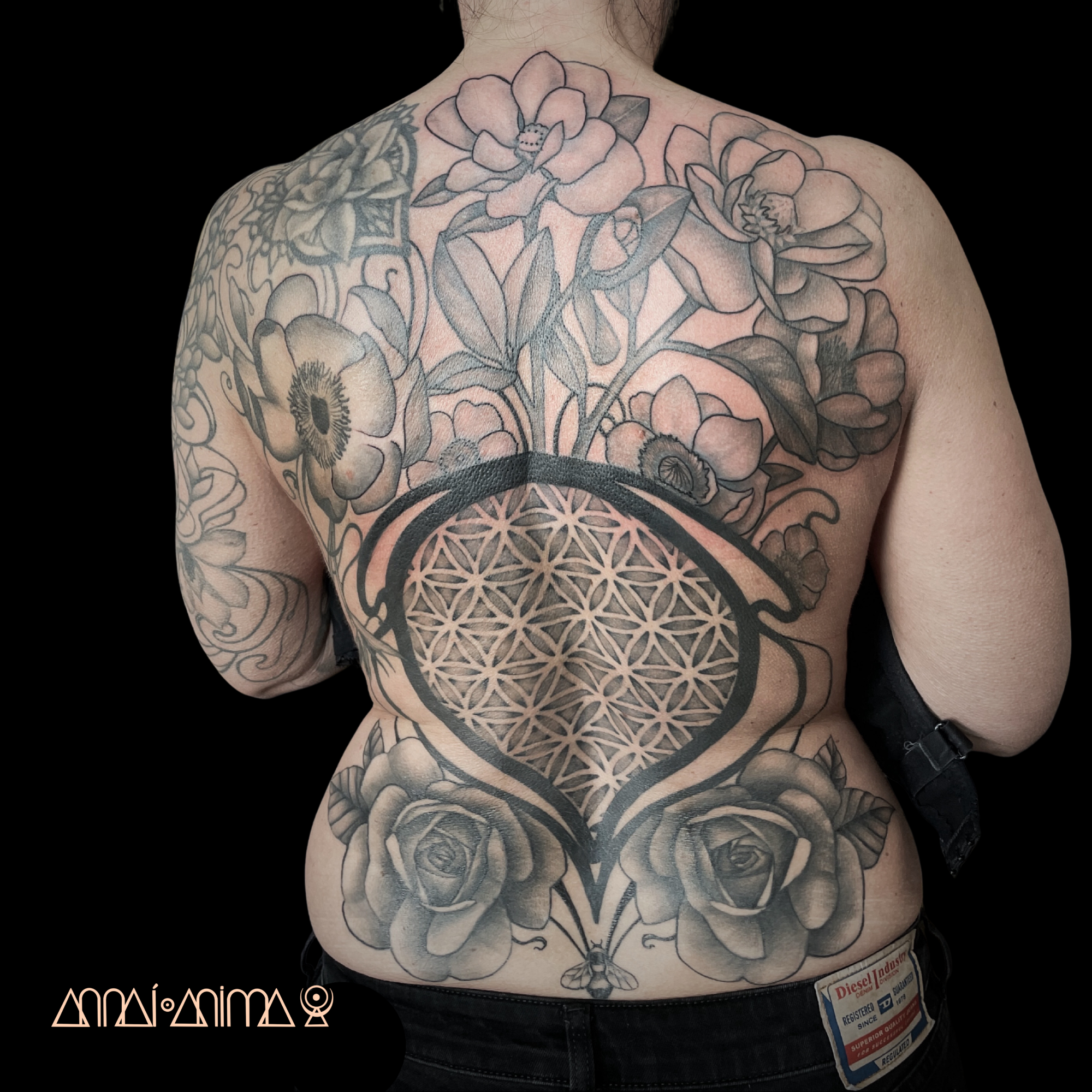Back of a person with extensive black and gray floral tattoos, including roses and anemones, and a large geometric pattern resembling the Flower of Life with a bee at the bottom.