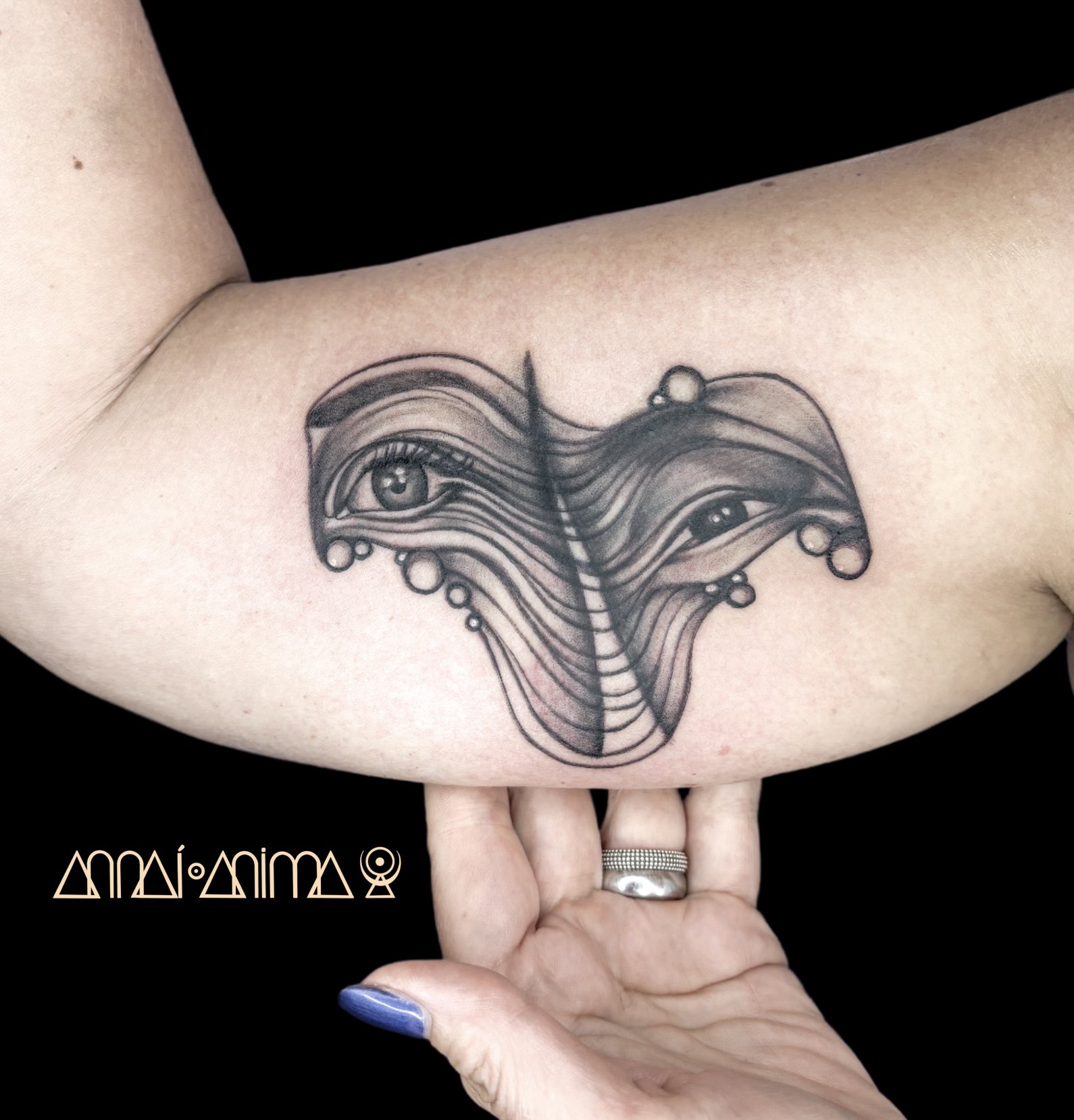 A black and gray tattoo of an abstract face with eyes, flowing lines, and circular details on a person's upper arm.