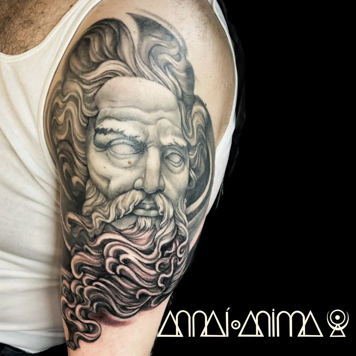 Black and gray tattoo of a bearded ancient Greek or Roman god or philosopher on upper arm, with stylized symbols or text below.